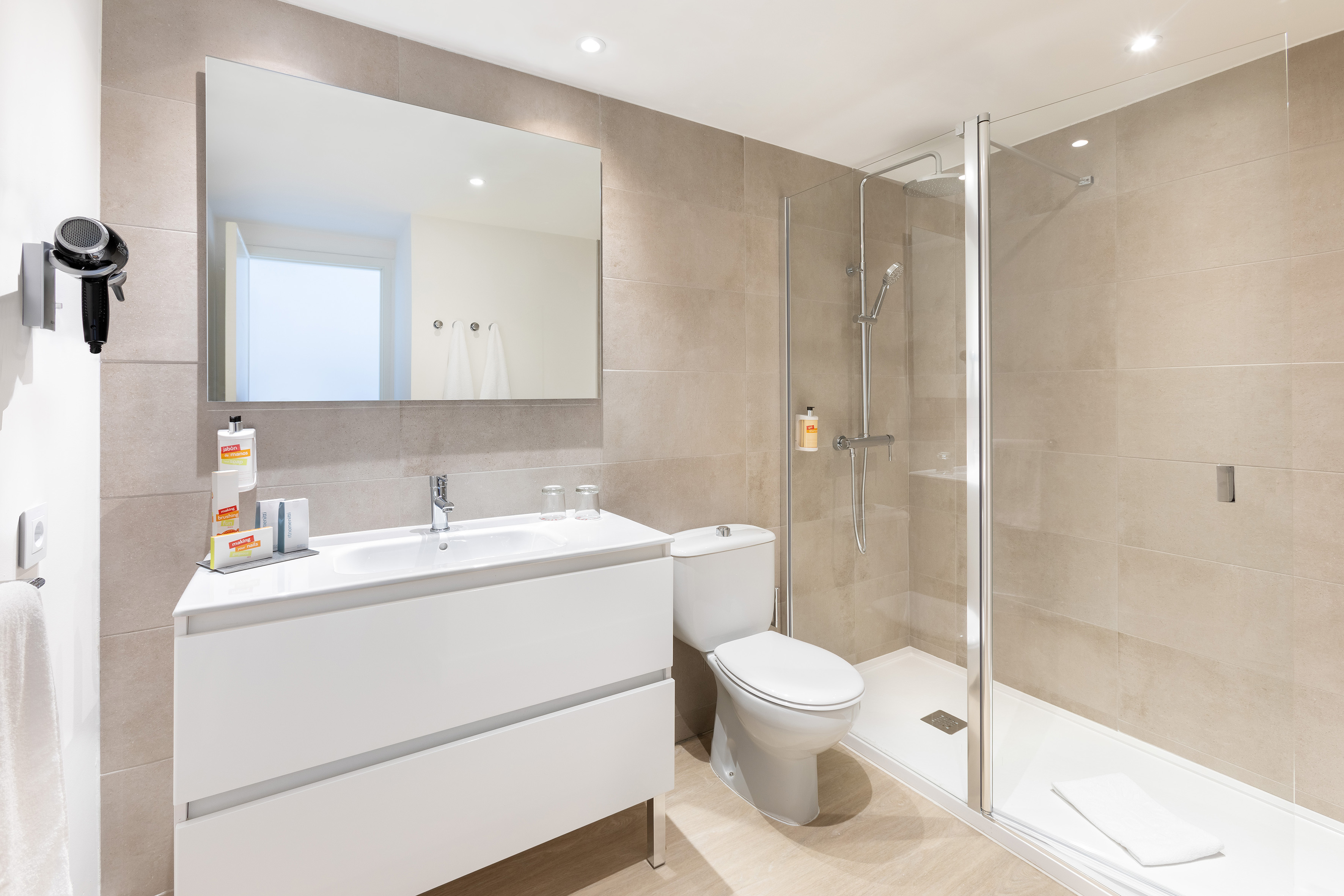 Modern bathroom with beige tiles, white vanity, mirror, shower, toilet, hairdryer.