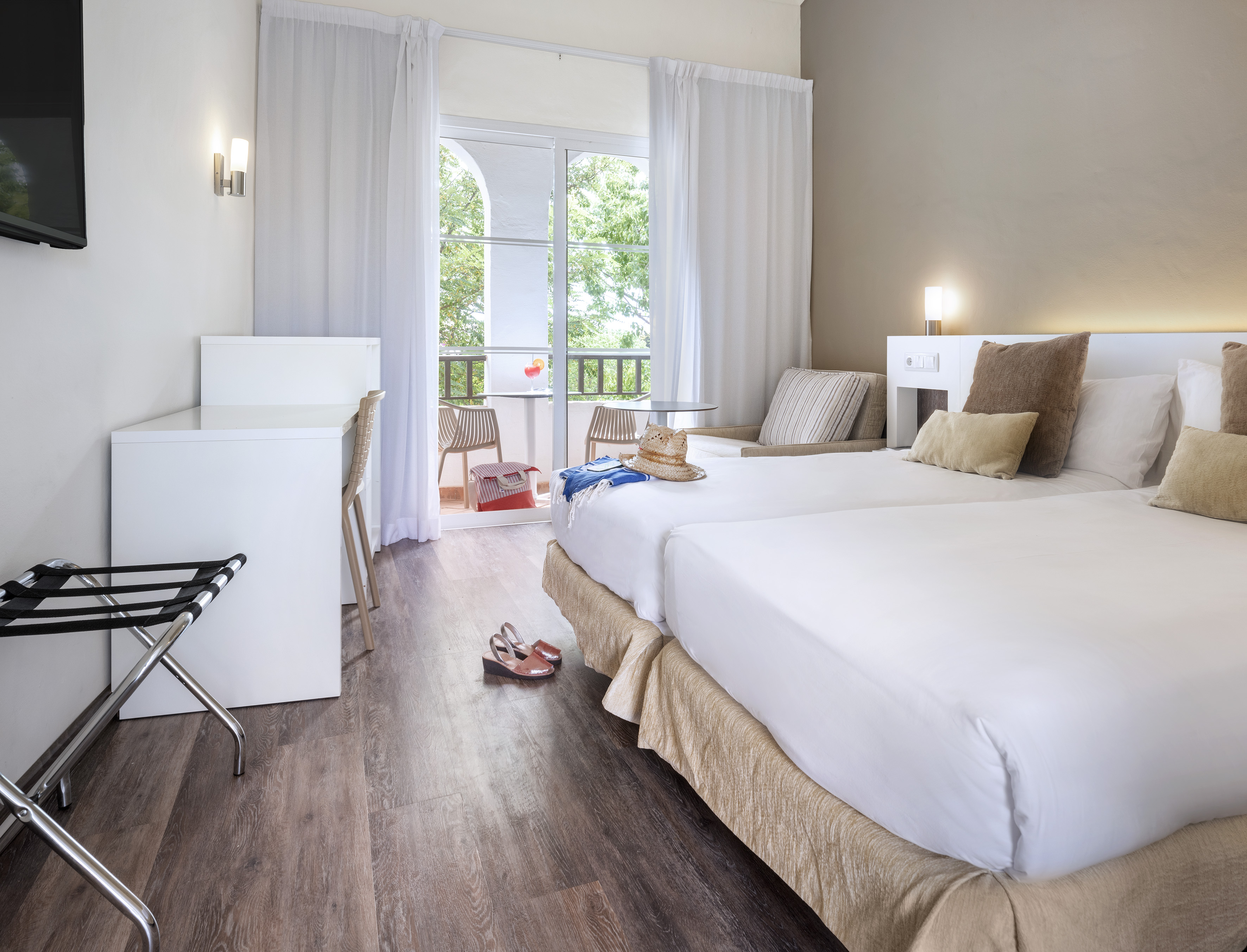 Hotel room with twin beds, wooden floor, and a sunlit balcony.