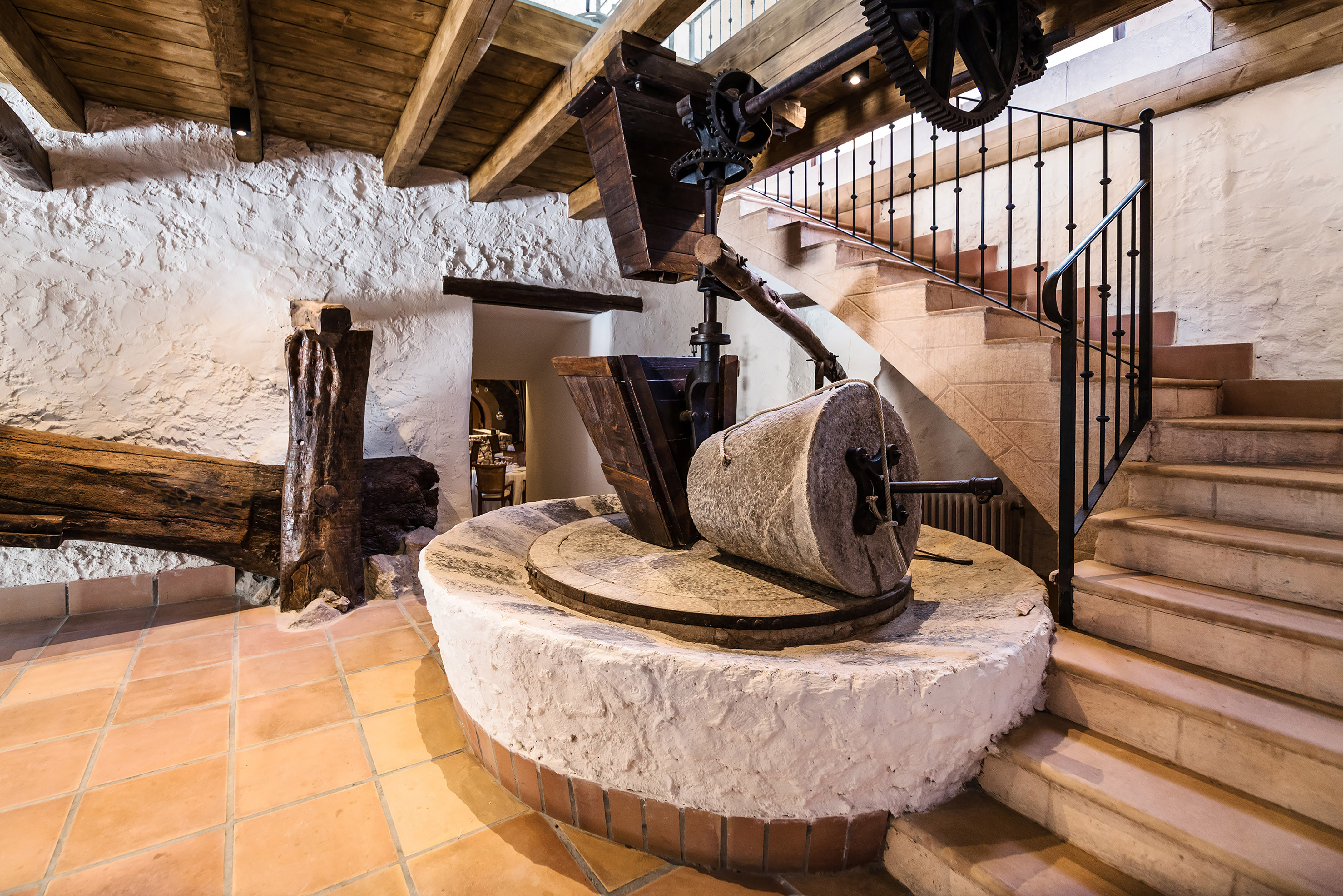Rustic stone olive press, wooden gears, terracotta floor, and stone stairs.
