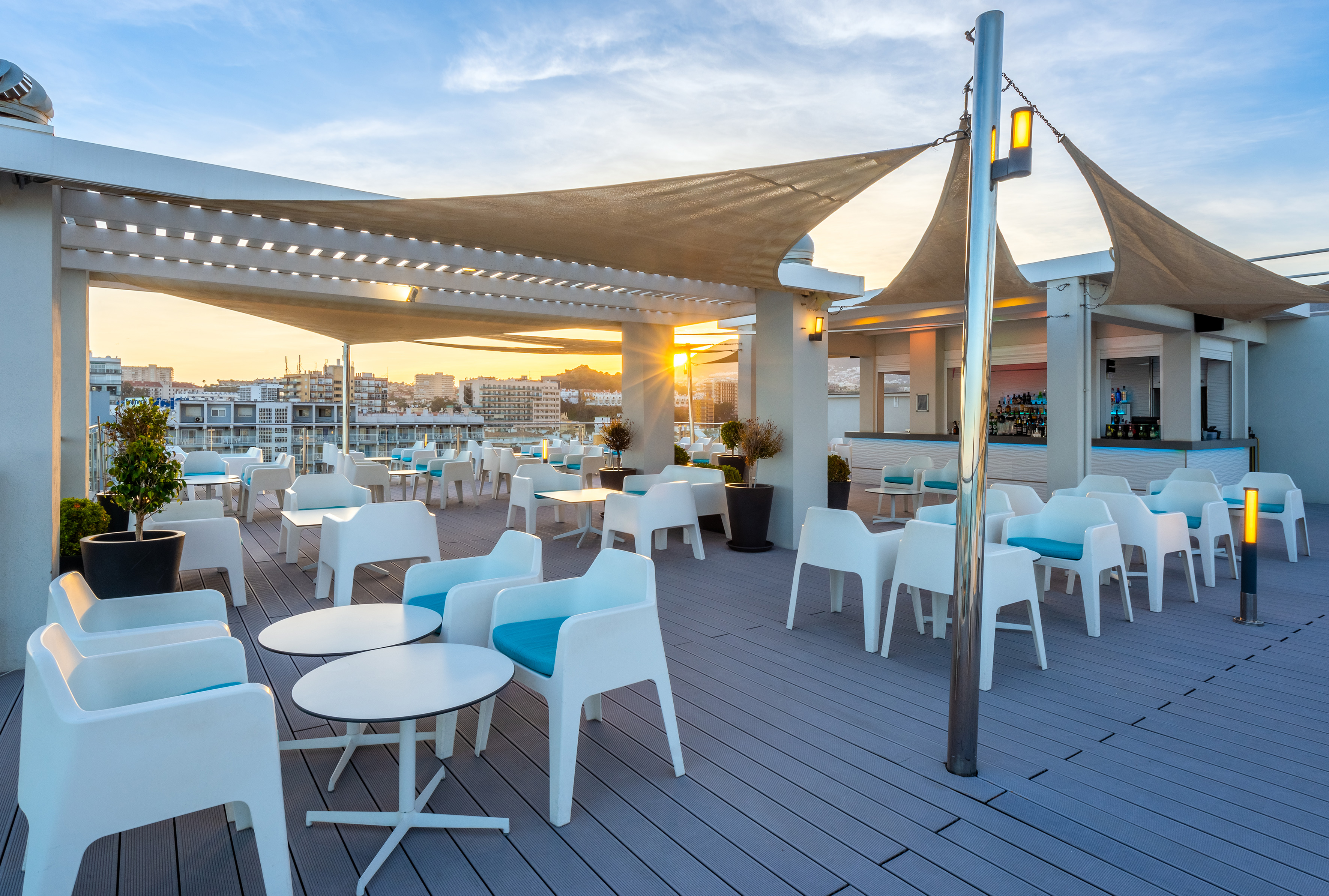 Rooftop bar with white chairs, blue cushions, shade sails, sunset city view.
