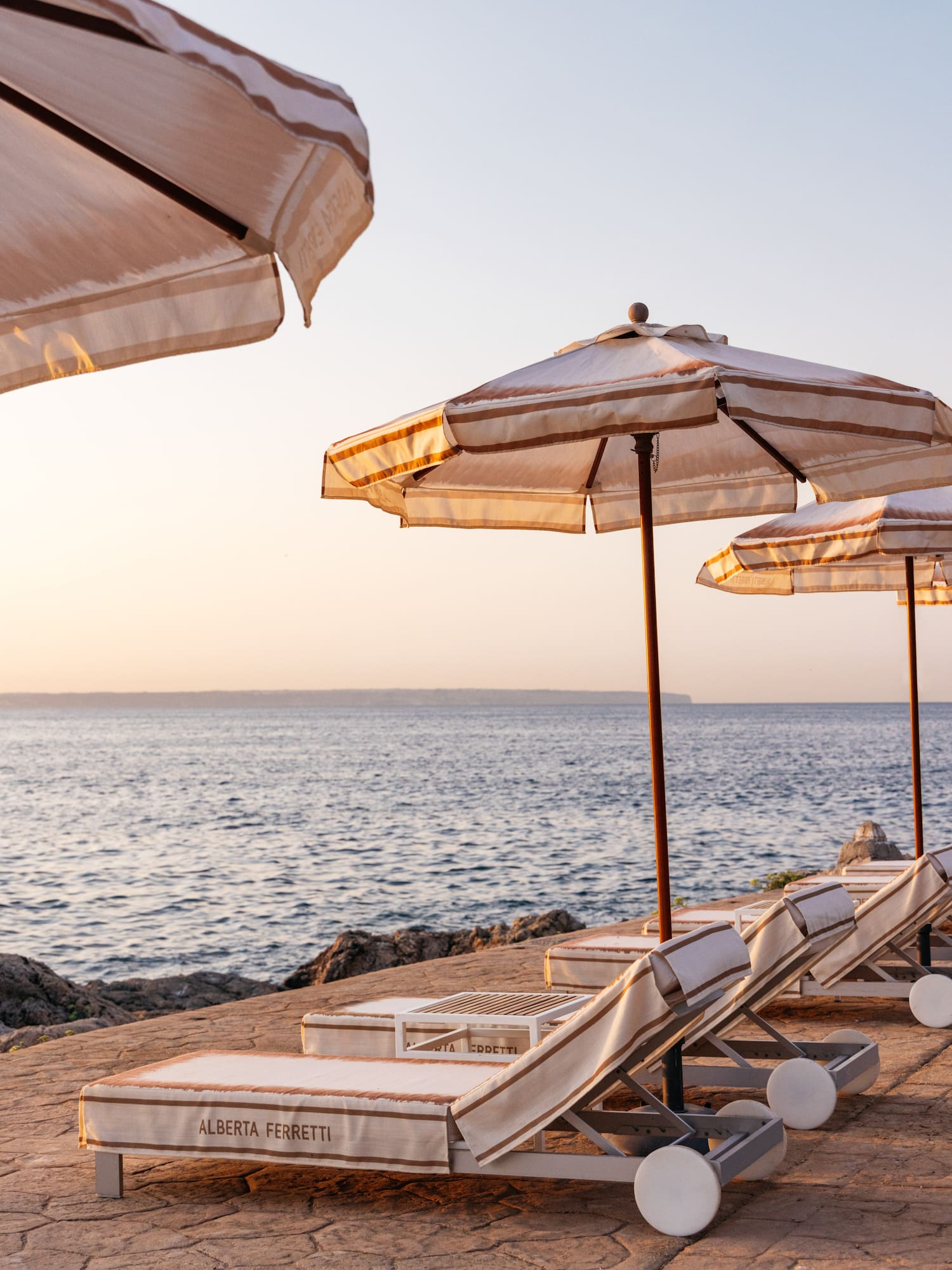 Alberta Ferretti luxury lounge chairs and striped umbrellas by the ocean at sunset.