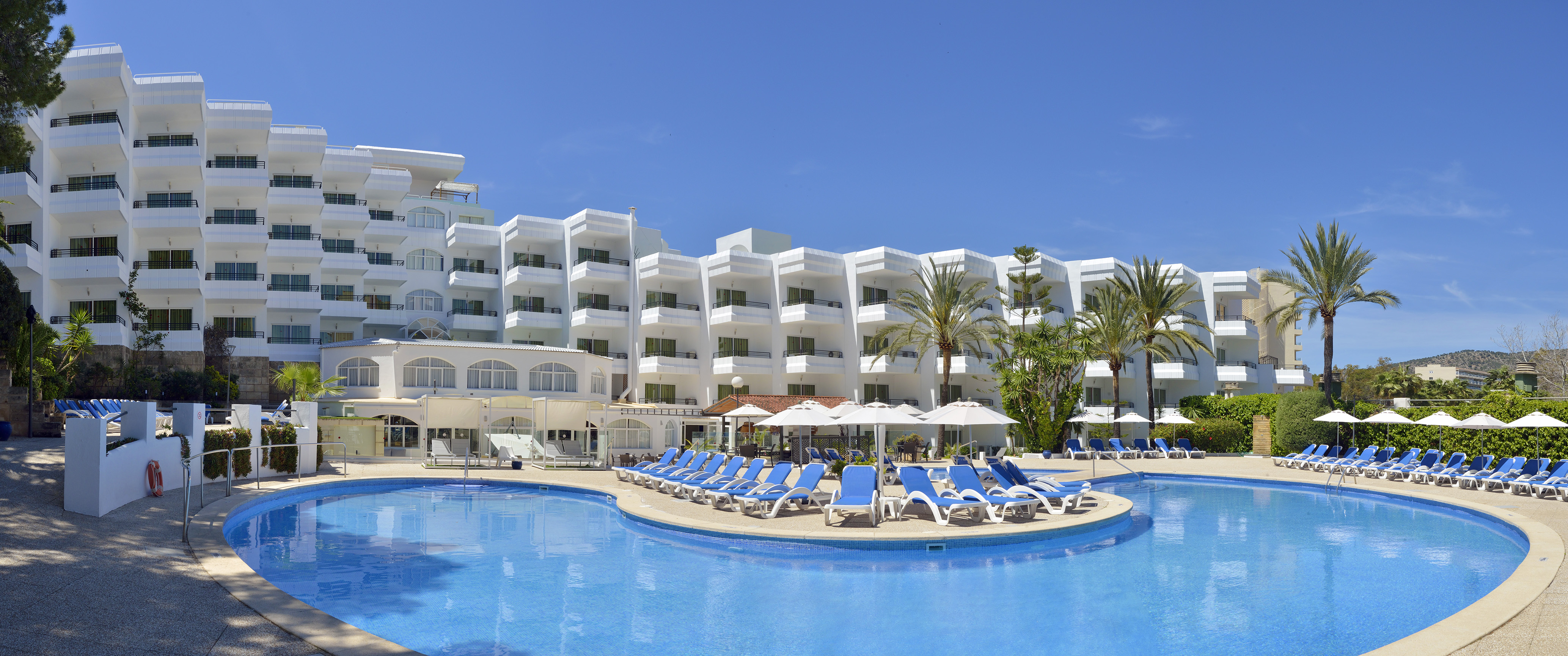 Sunny resort hotel with large outdoor pool, blue lounge chairs, and palm trees.