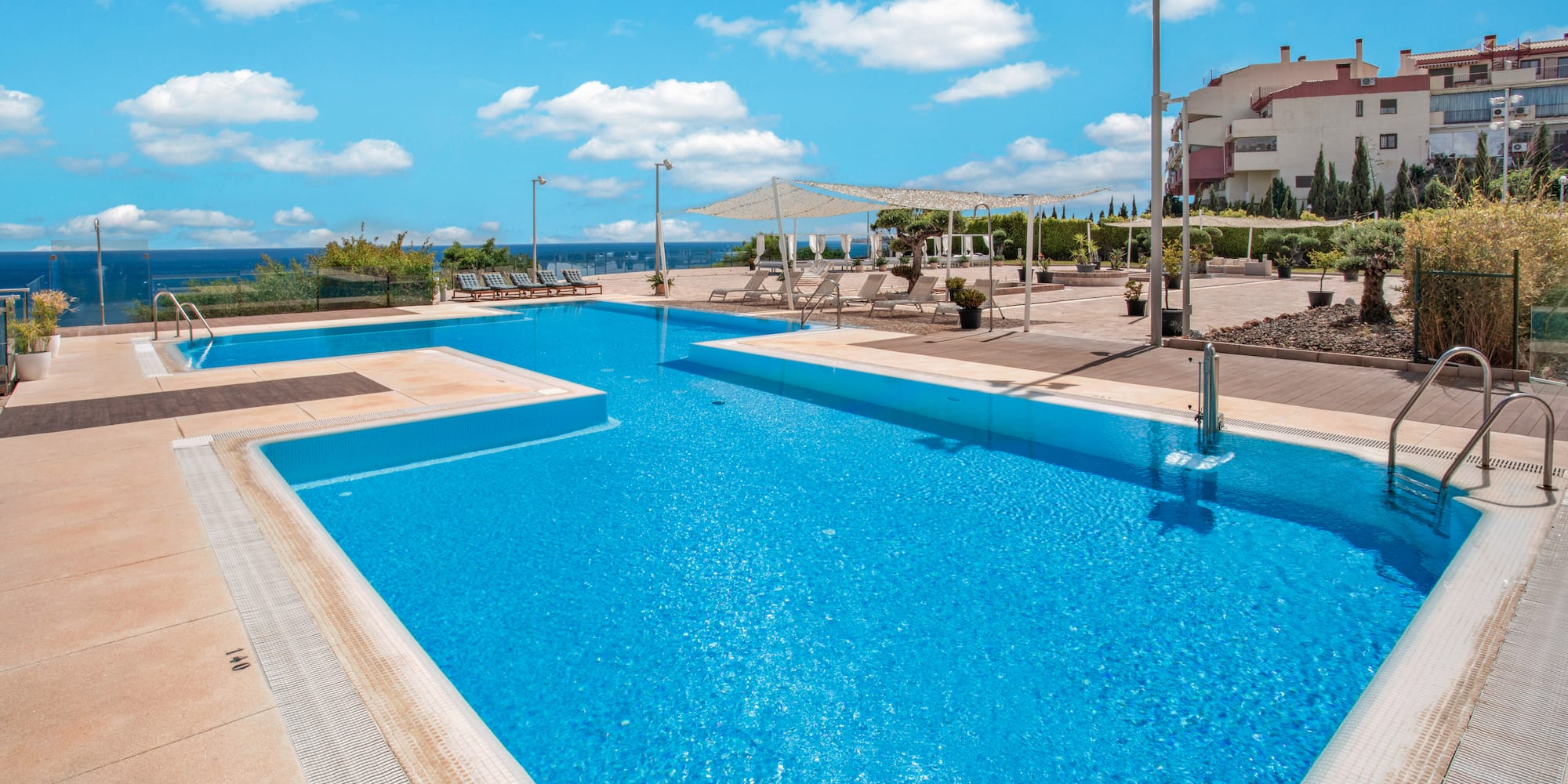 Sunny resort swimming pools with ocean view, sunbeds, and apartment buildings.
