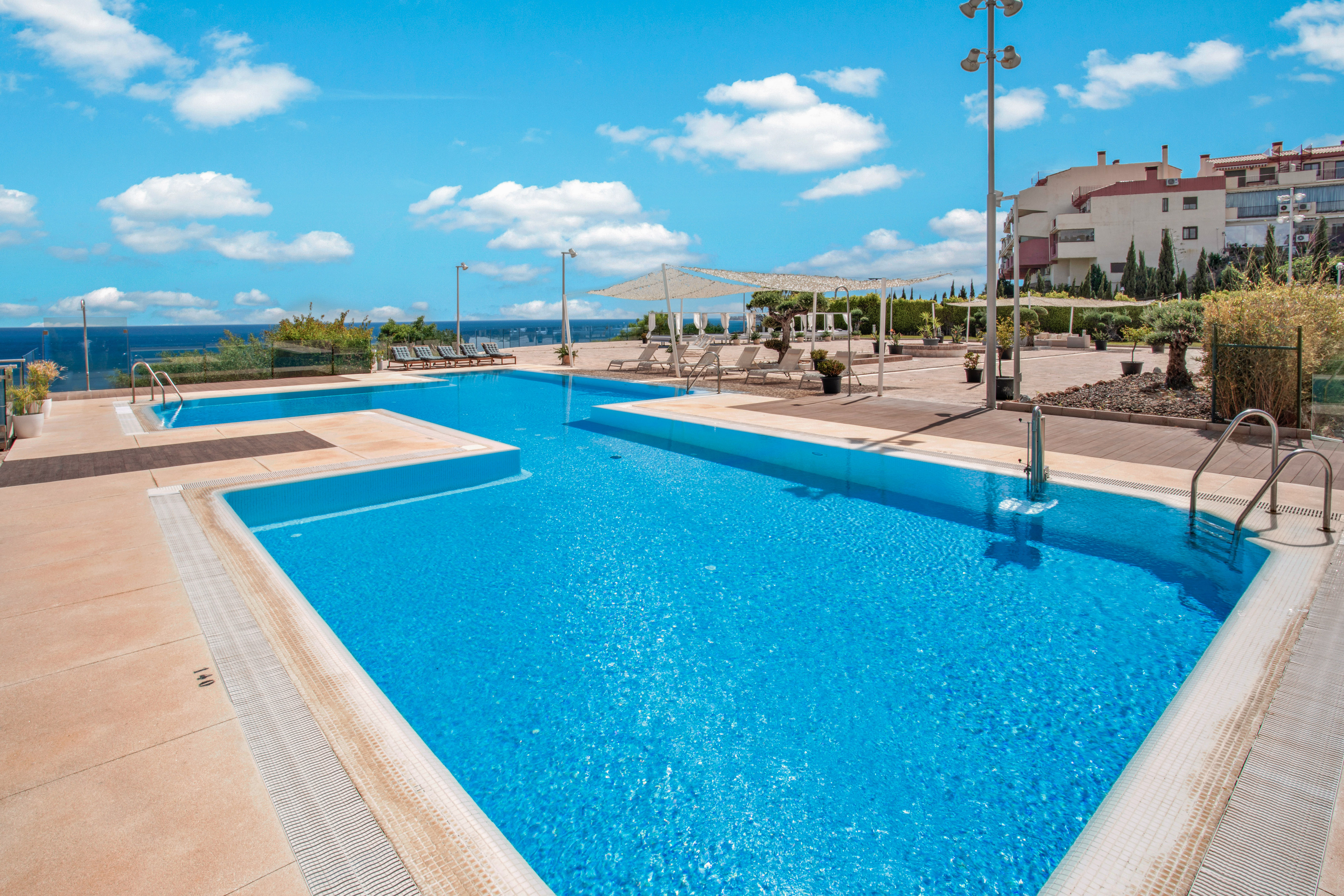 Sunny resort swimming pools with ocean view, sunbeds, and apartment buildings.