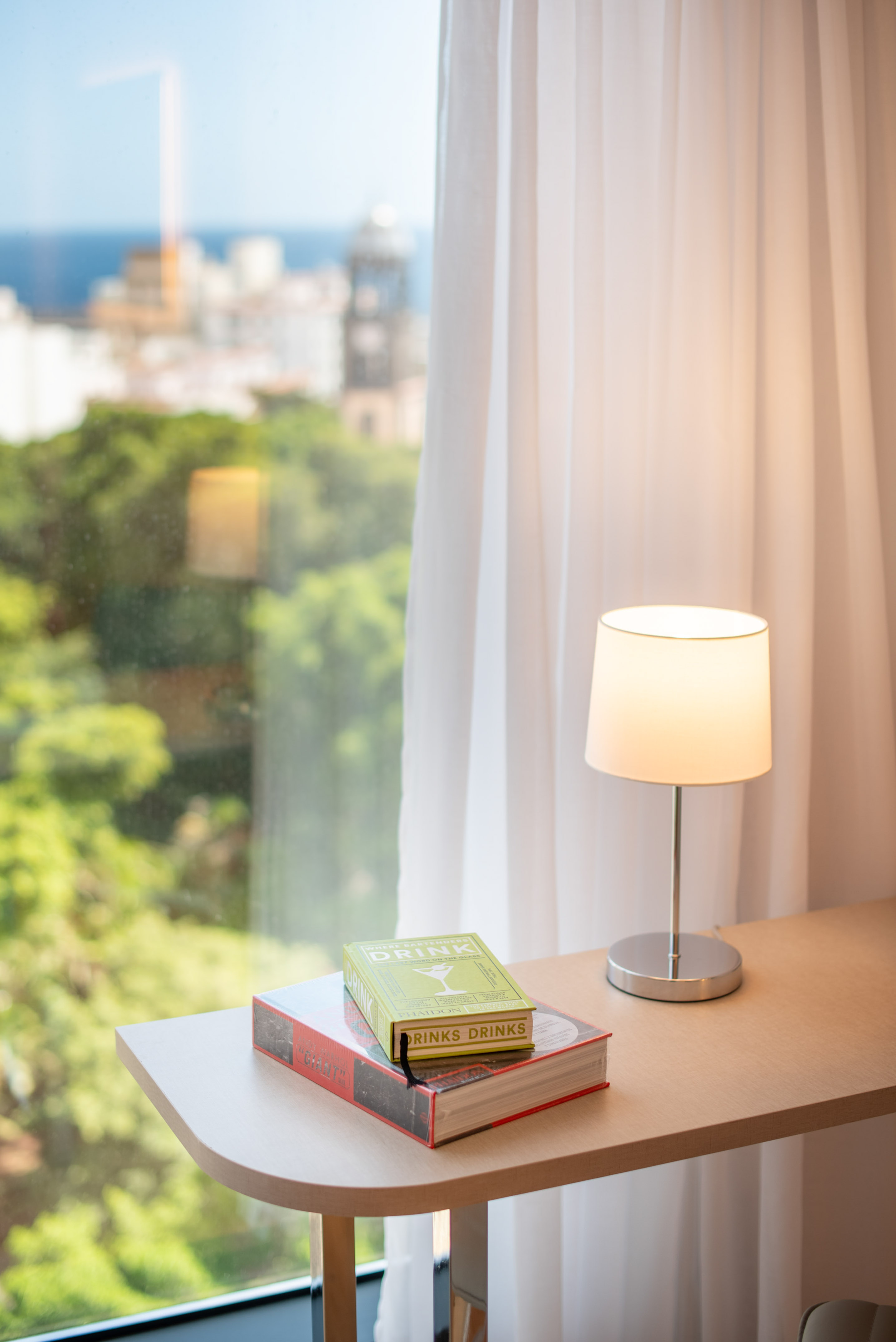 Desk with 'Drink' books, lamp, sheer curtain, overlooking city, trees, ocean.