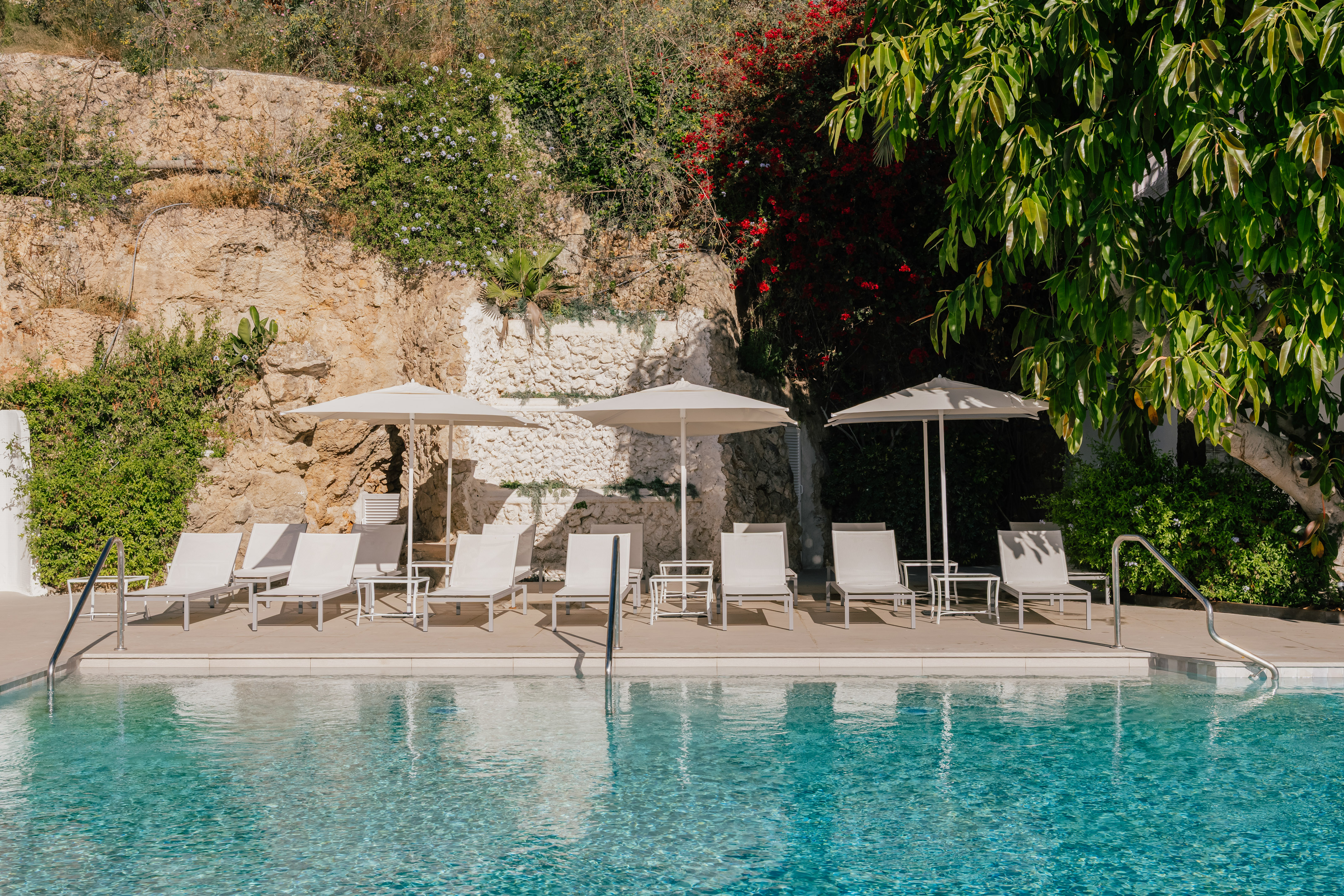 Sunny pool, white lounge chairs, umbrellas, and lush green rocky landscape.