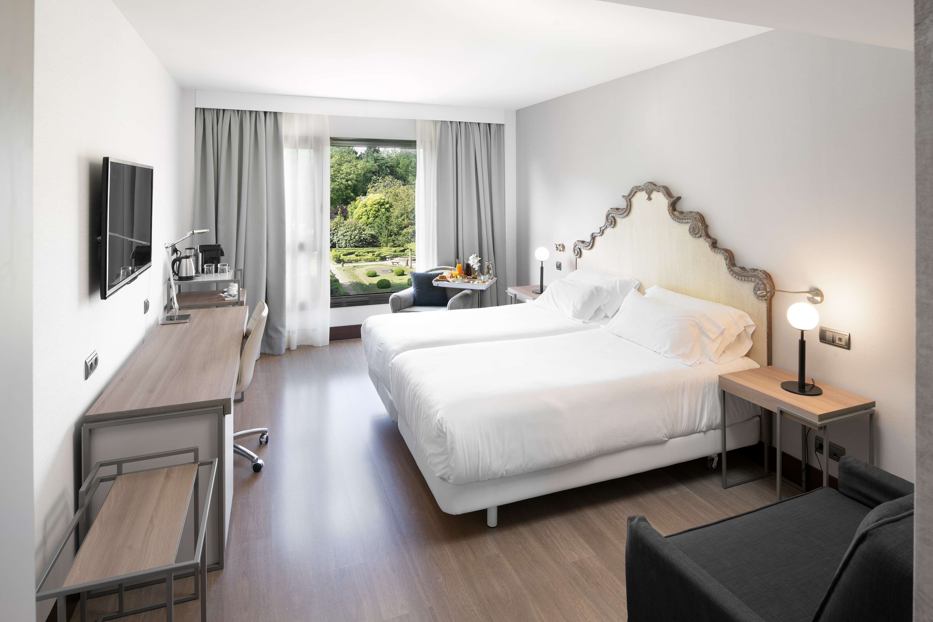 Bright hotel room with two beds, desk, ornate headboard, and garden view.
