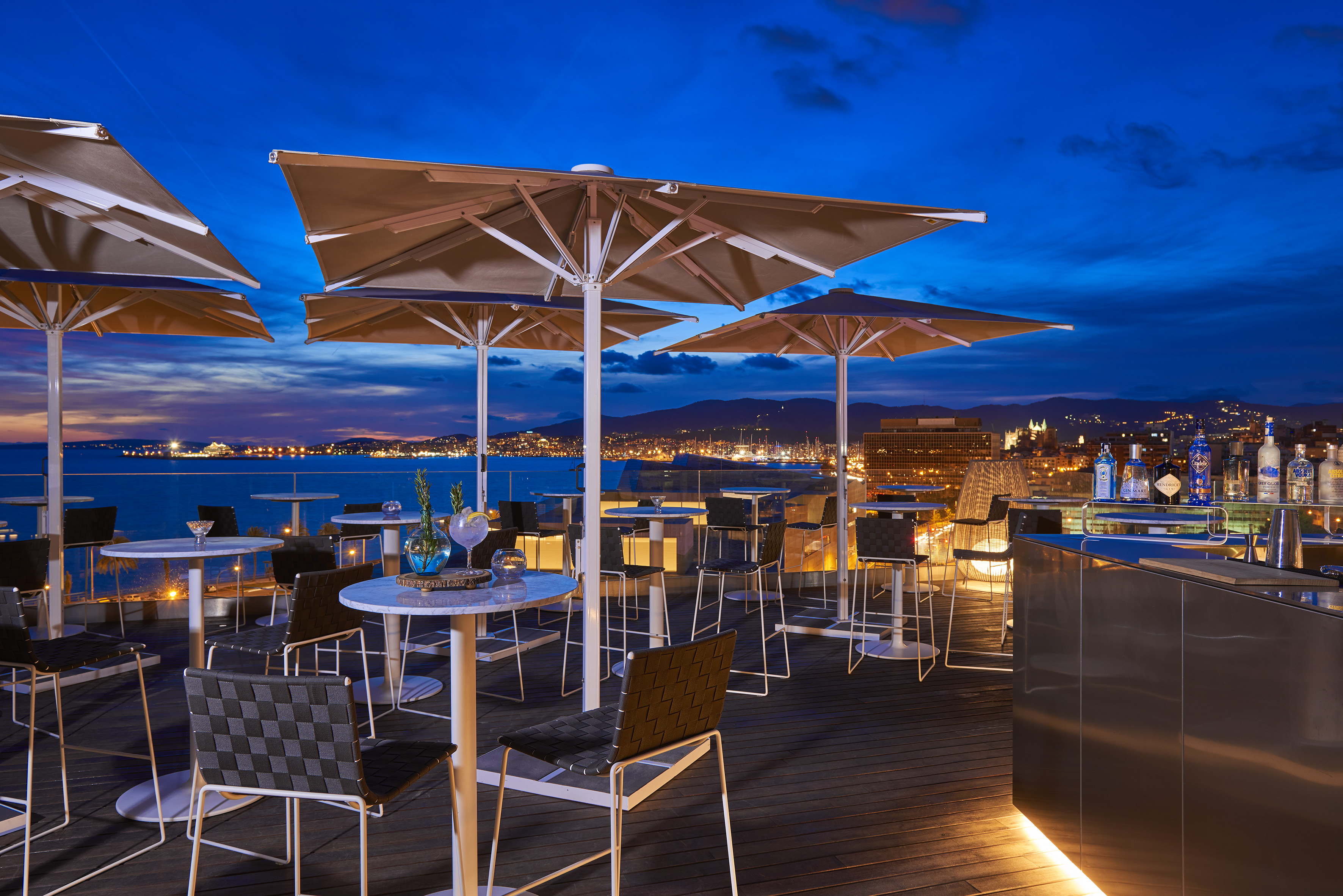 Rooftop bar terrace, evening city and sea view, outdoor seating.
