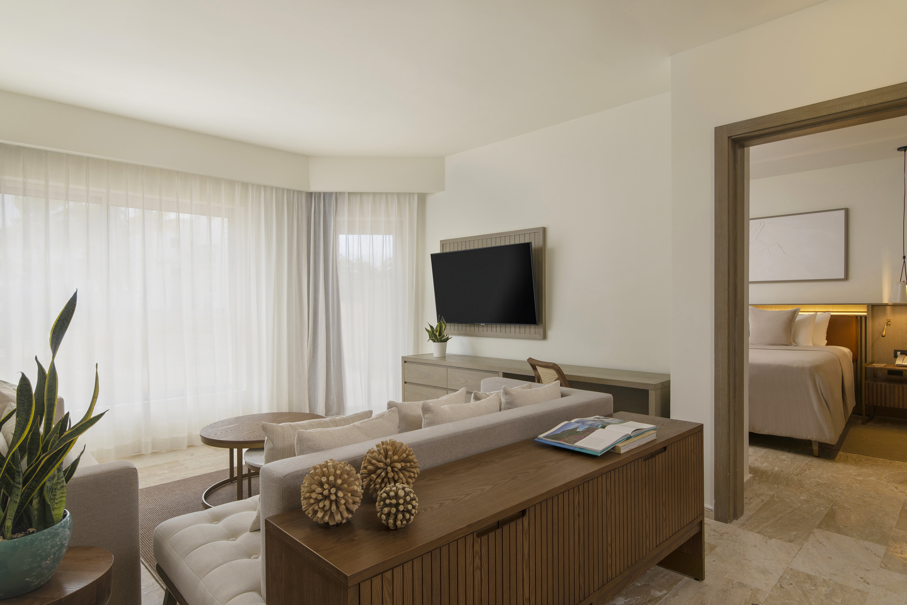 Modern hotel living room with sofa, mounted TV, and adjoining bedroom.
