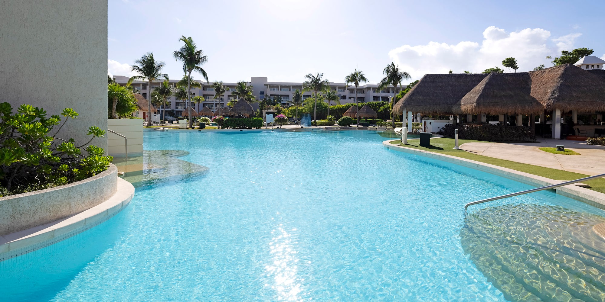 Sunny resort pool with palm trees, hotel buildings, and thatched poolside huts.