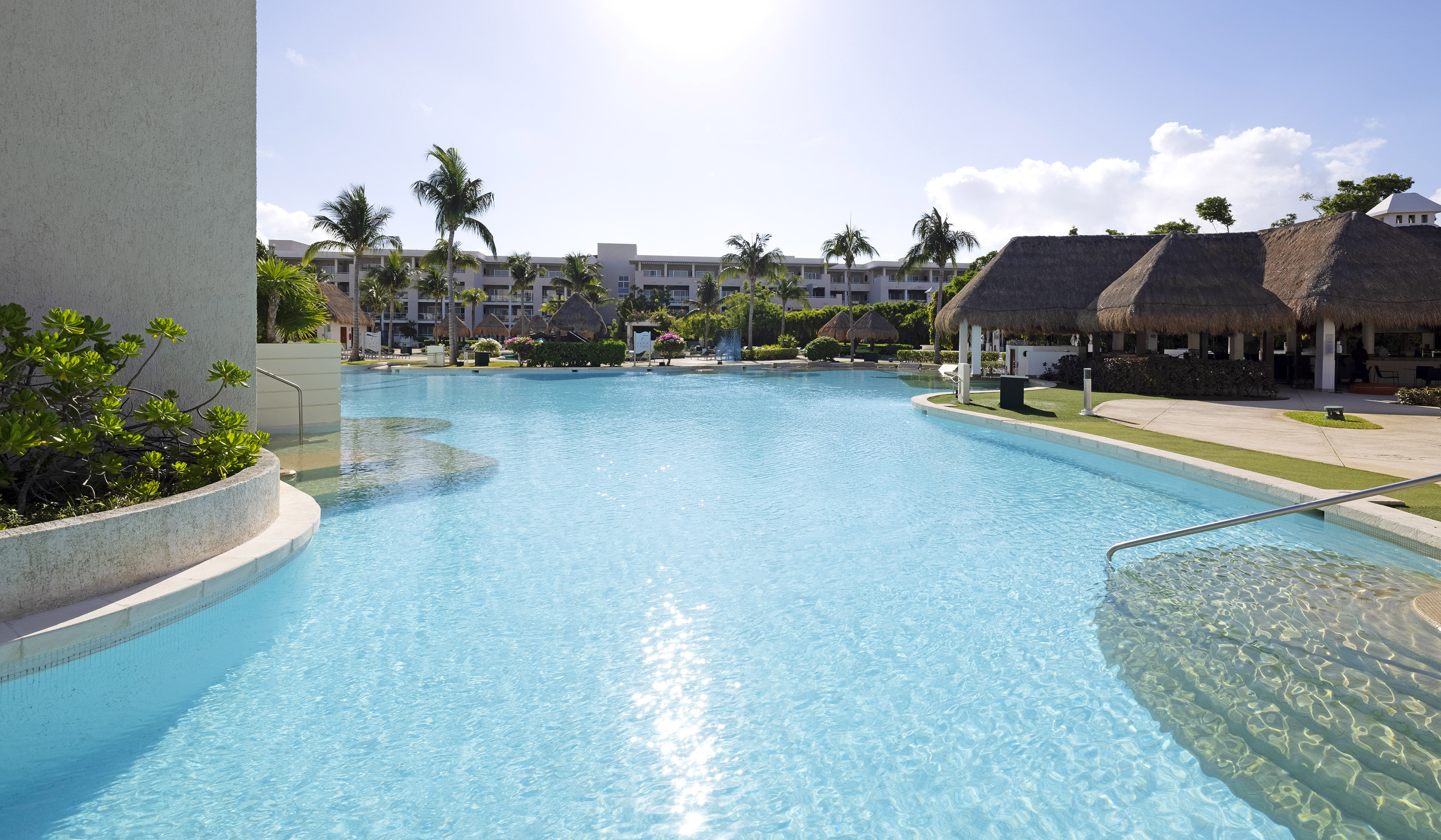Sunny resort pool with palm trees, hotel buildings, and thatched poolside huts.