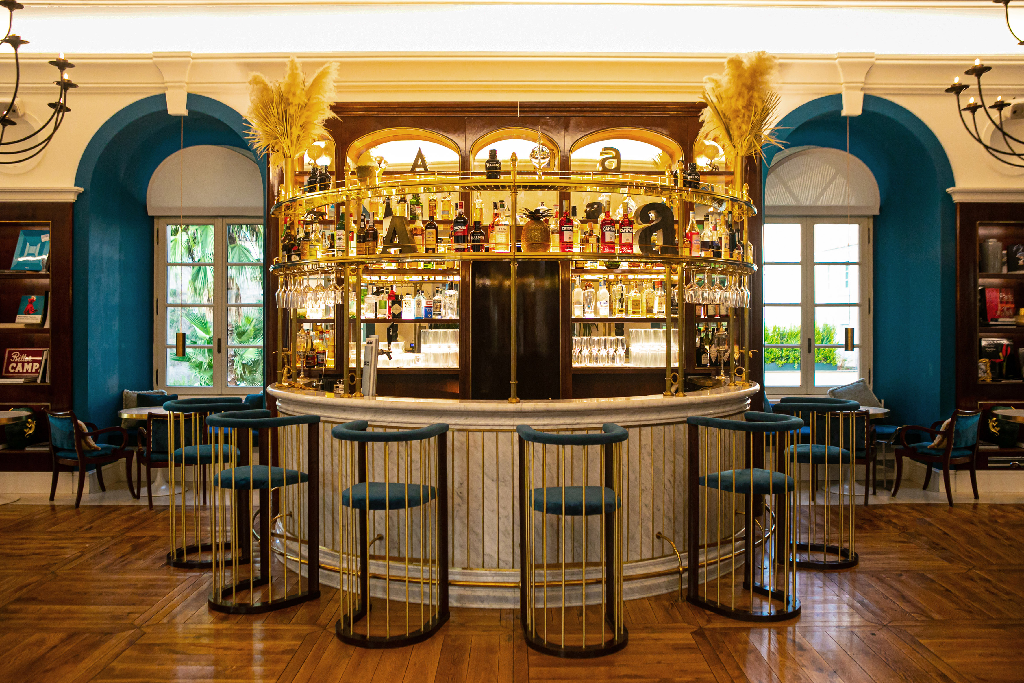 Elegant circular brass bar with marble, teal velvet stools, arched windows.