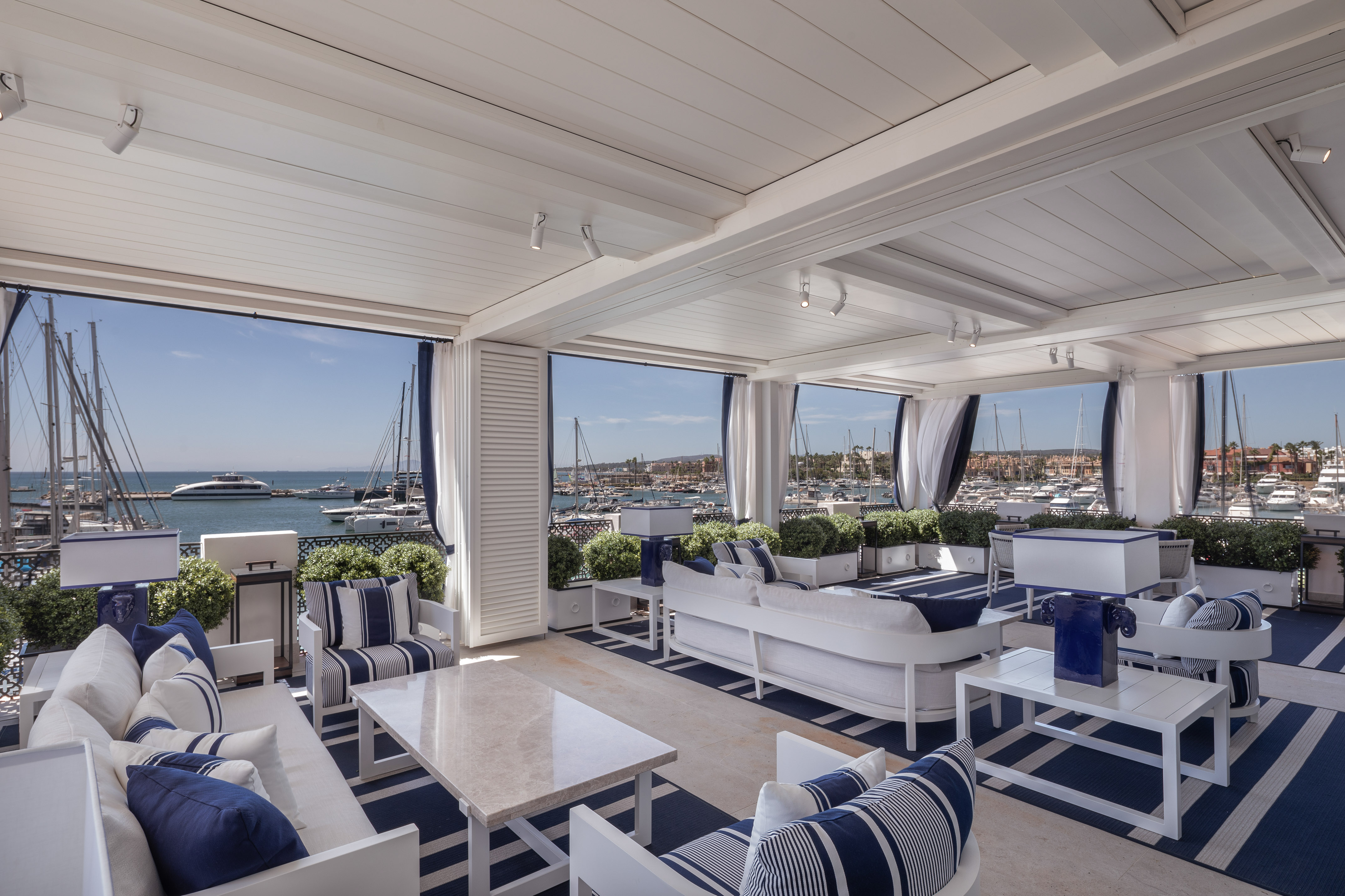 Outdoor patio with white furniture, blue accents, overlooking a sunny marina with yachts.