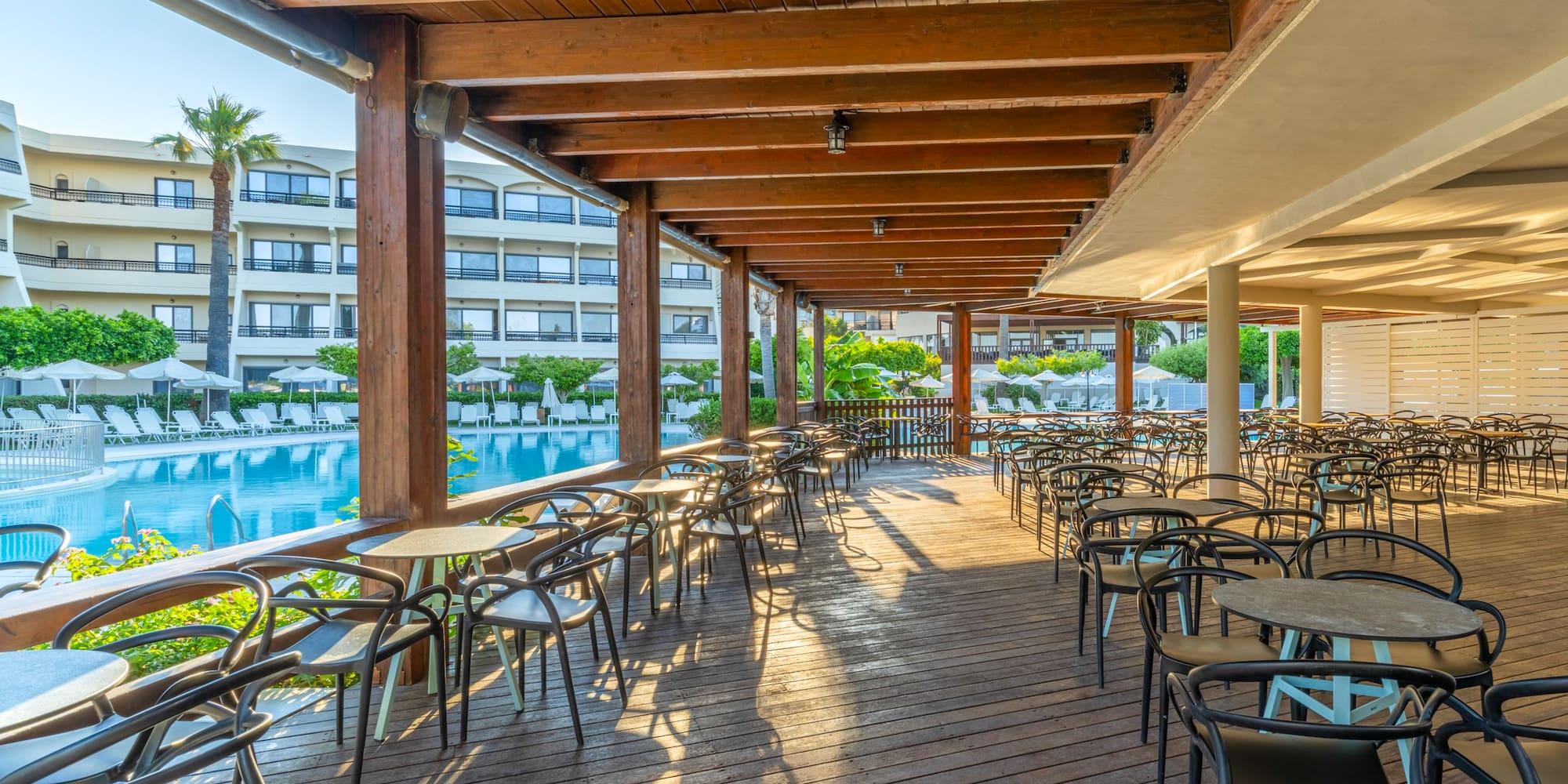 Sunny outdoor restaurant patio with wooden deck, tables, and swimming pool.