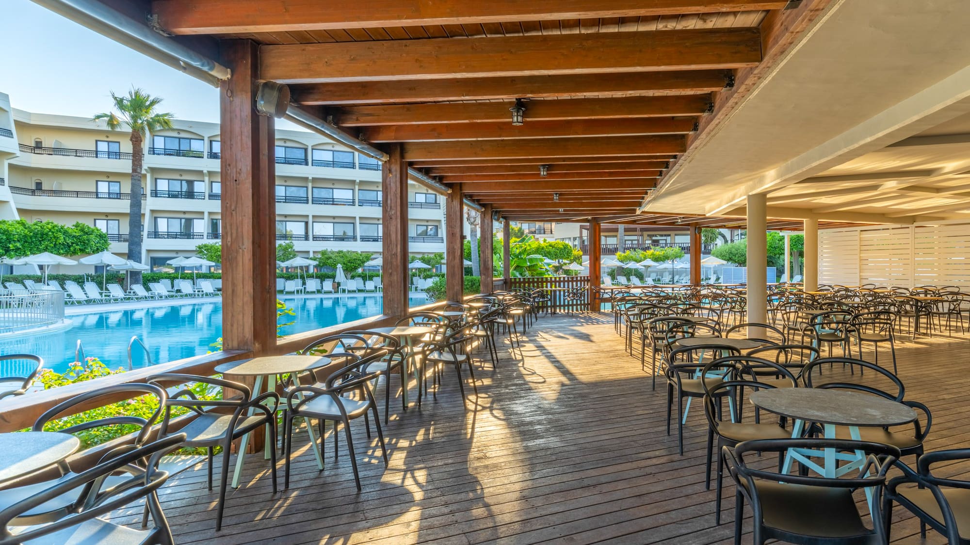 Sunny outdoor restaurant patio with wooden deck, tables, and swimming pool.