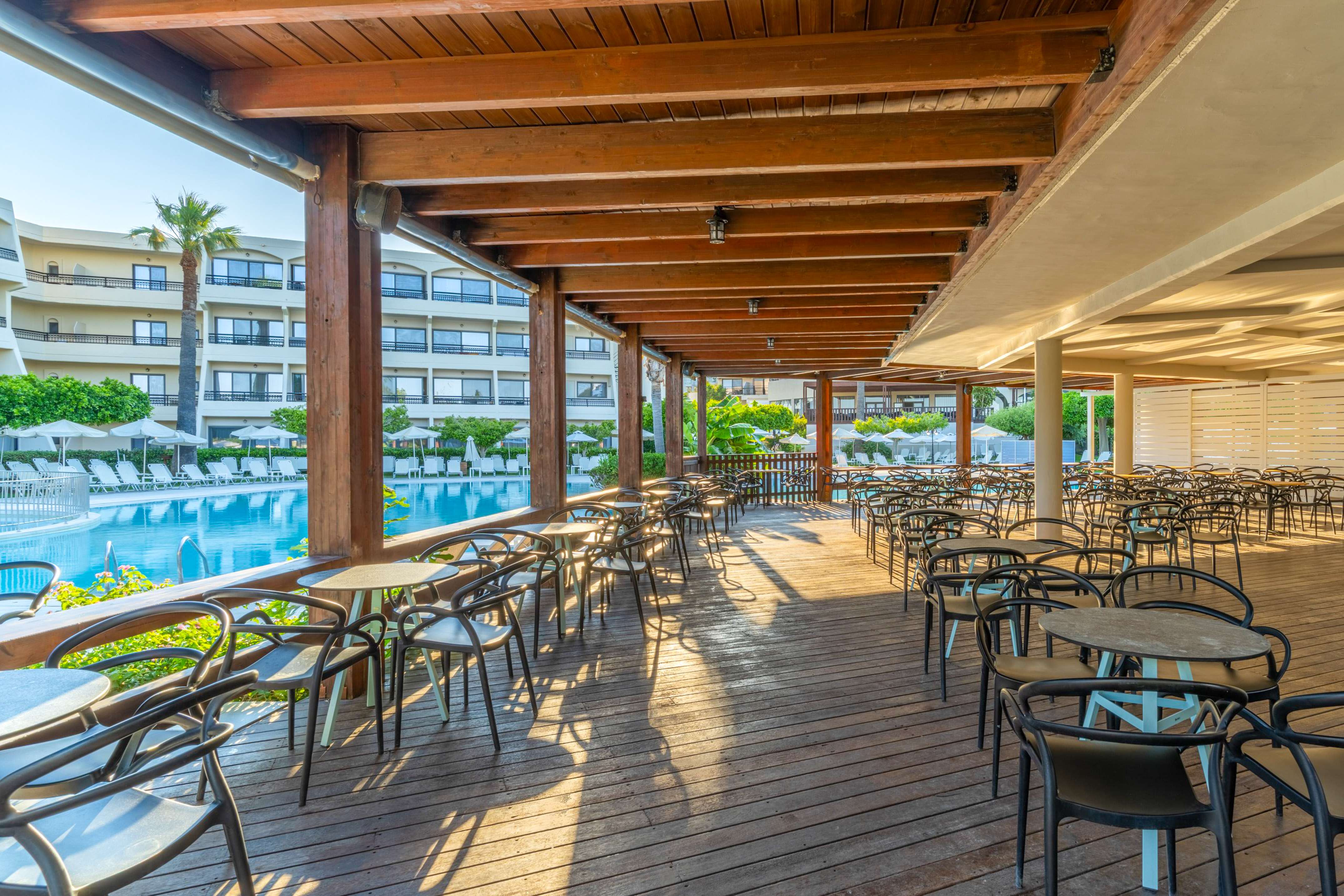 Sunny outdoor restaurant patio with wooden deck, tables, and swimming pool.