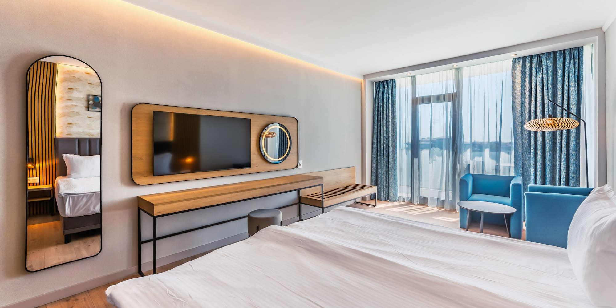 Modern hotel room featuring a bed, TV, blue armchairs, and large windows.