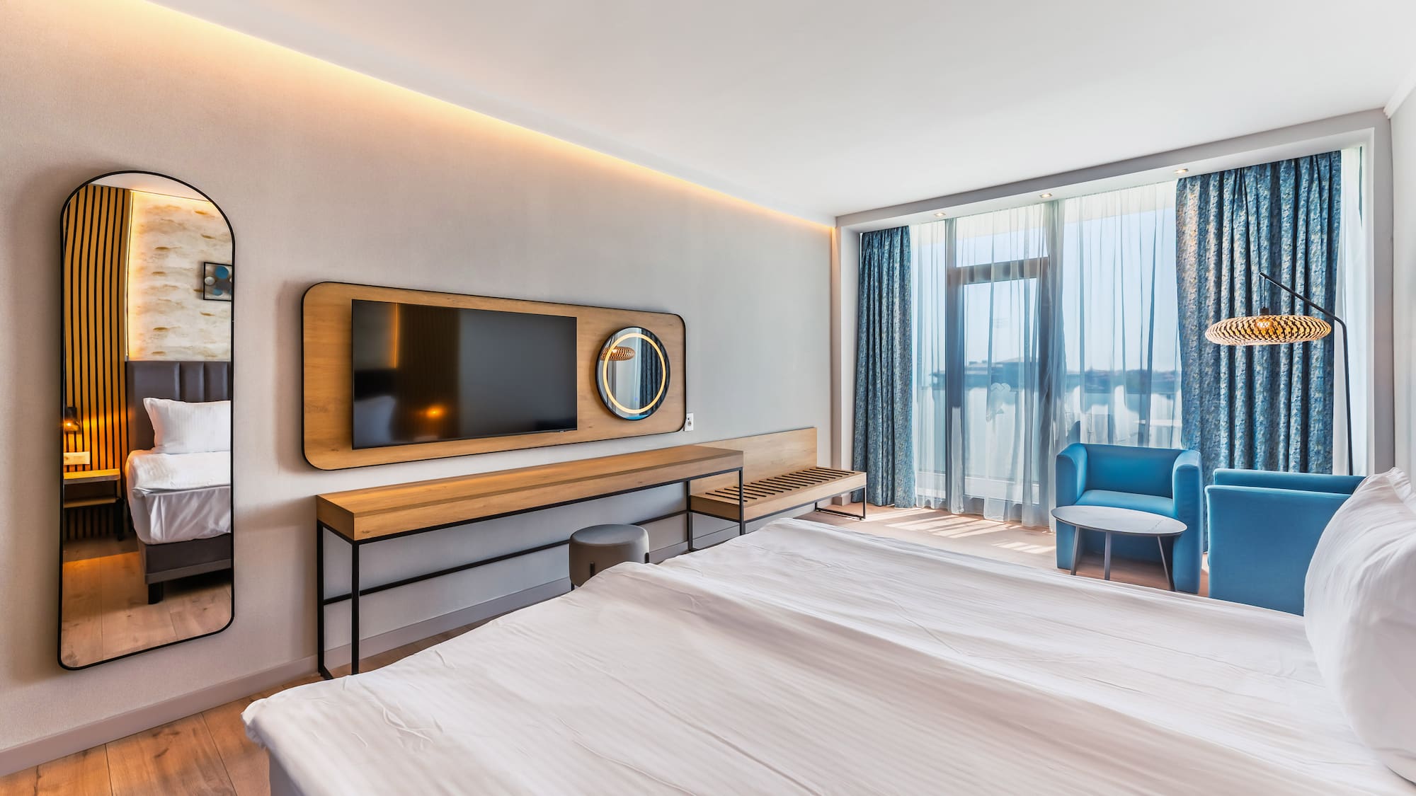 Modern hotel room featuring a bed, TV, blue armchairs, and large windows.