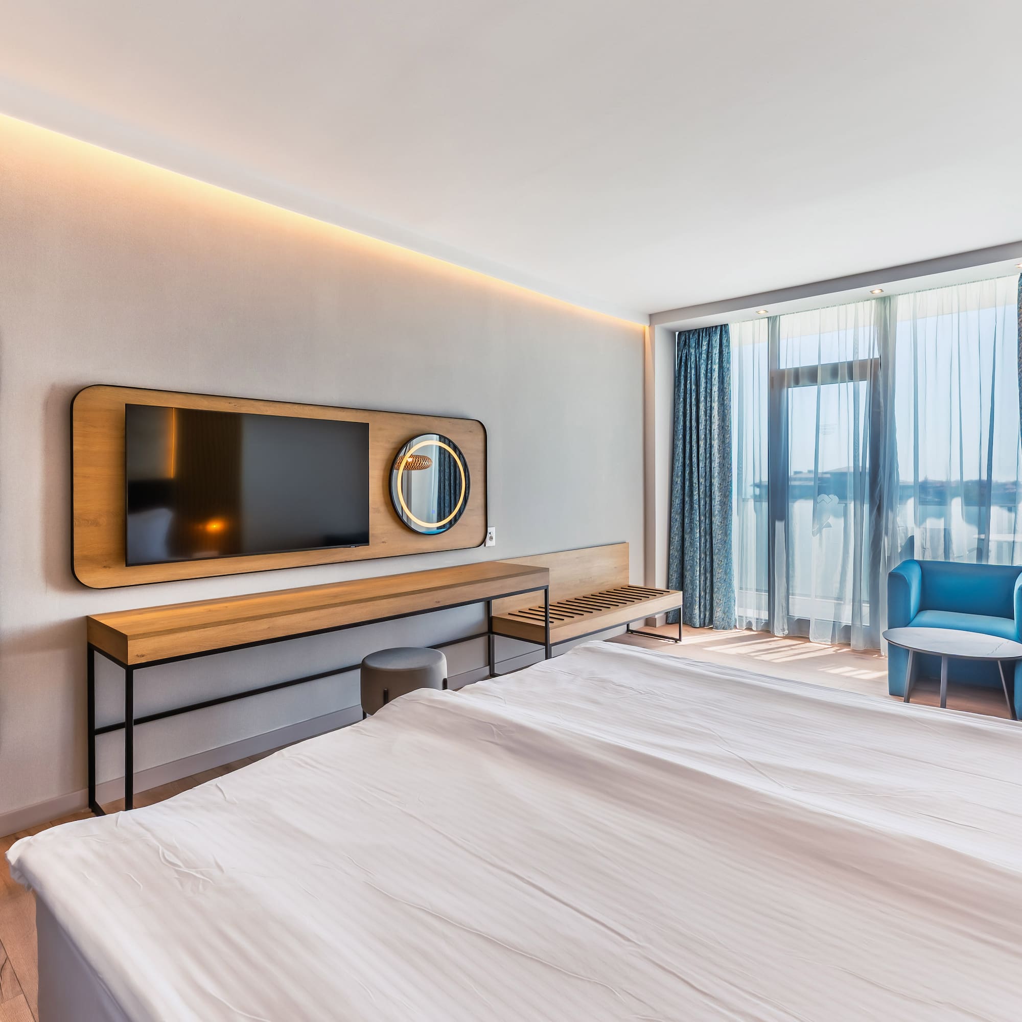 Modern hotel room featuring a bed, TV, blue armchairs, and large windows.