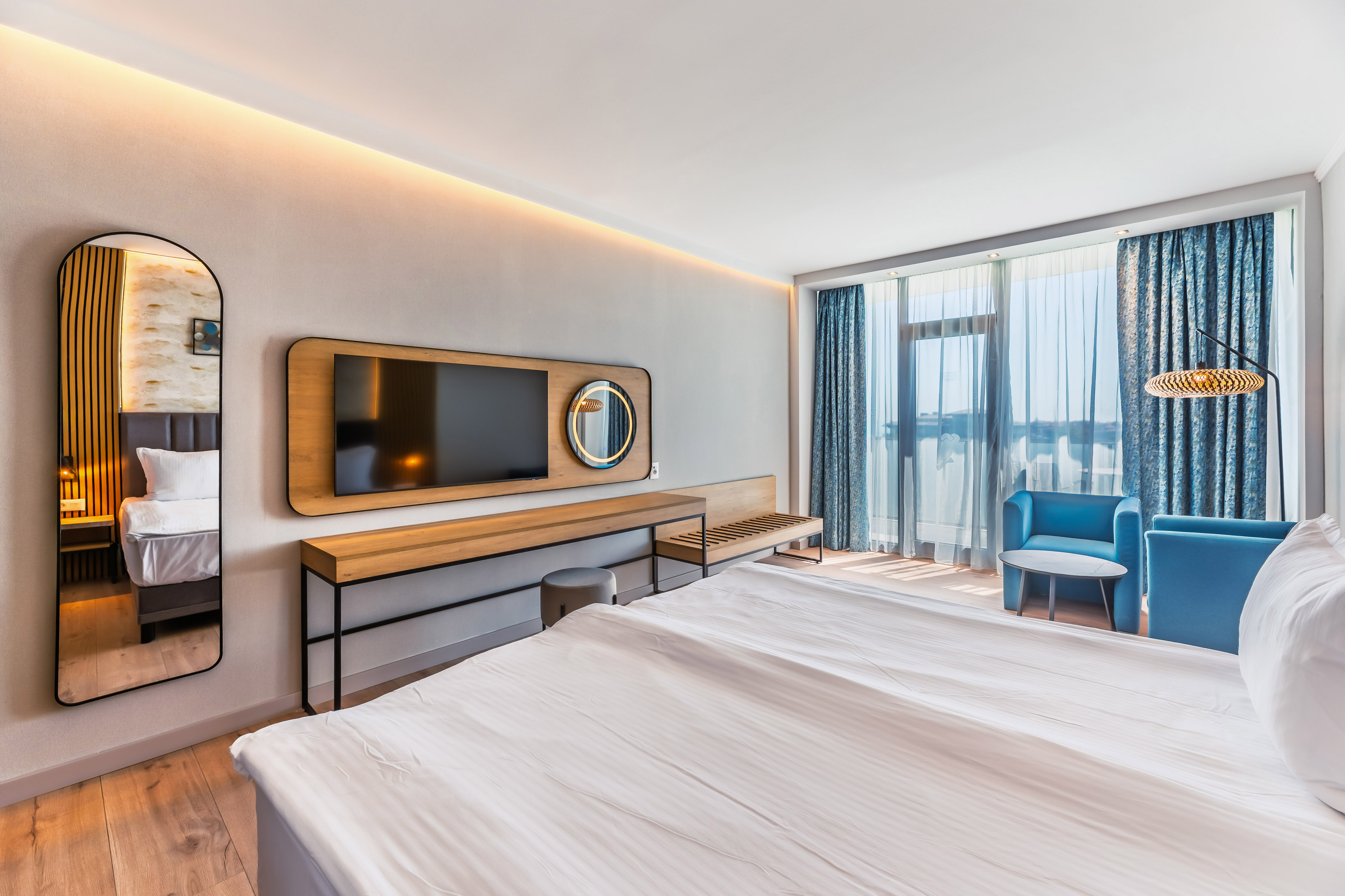 Modern hotel room featuring a bed, TV, blue armchairs, and large windows.