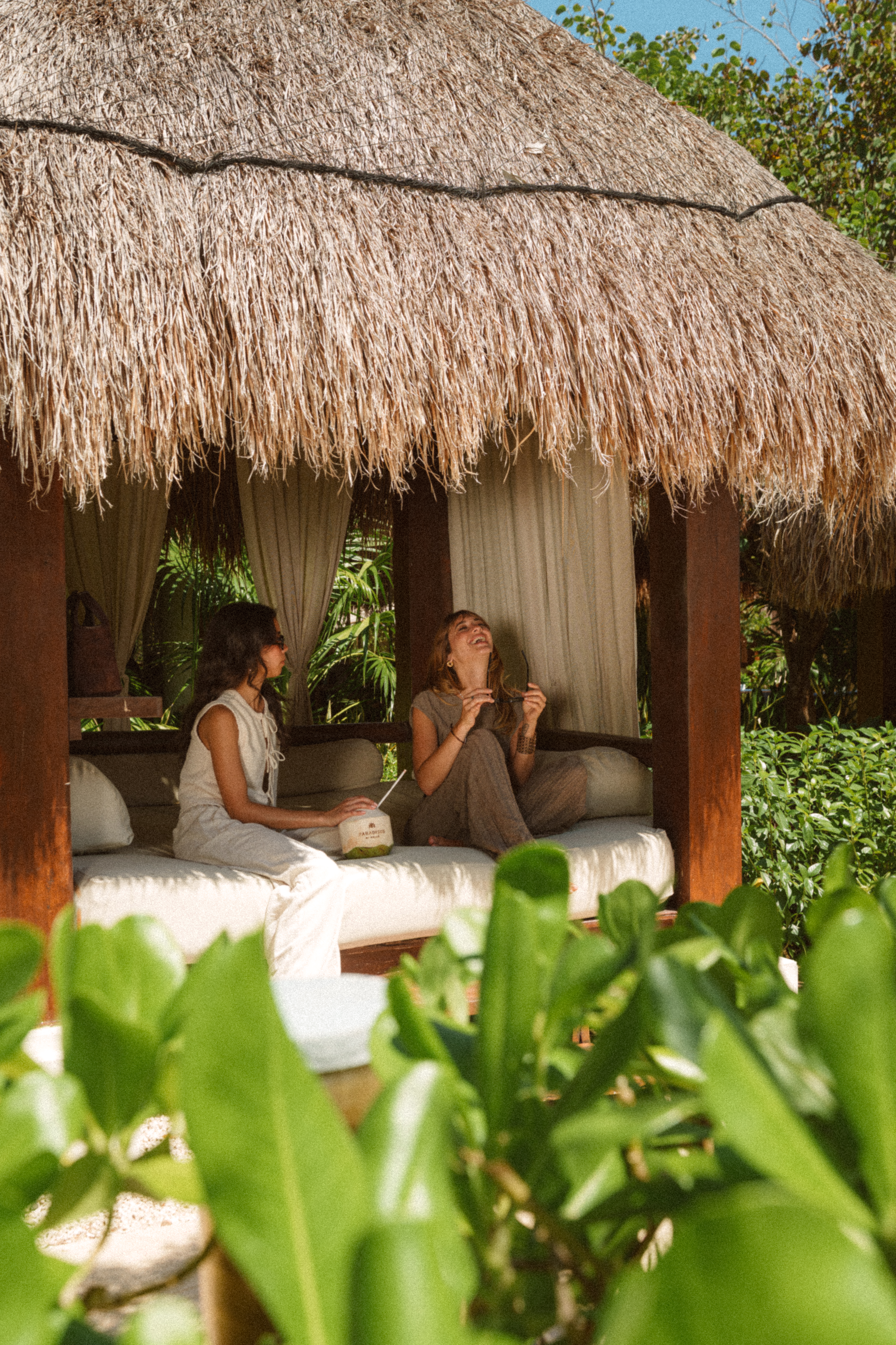 Women relaxing and laughing in a tropical thatched cabana with a coconut.
