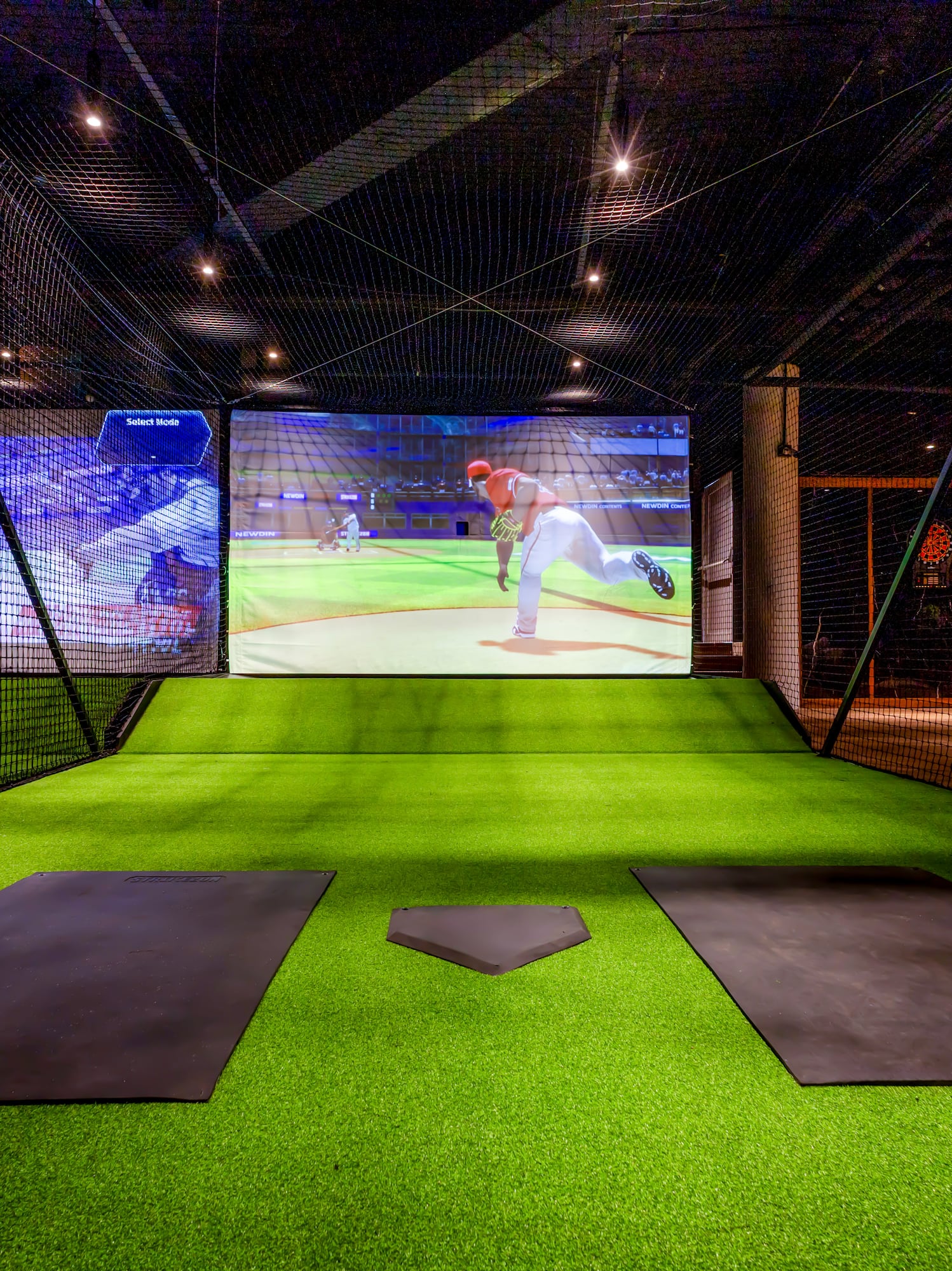 Simulador de béisbol virtual con pantalla, césped artificial y home plate en jaula de bateo.