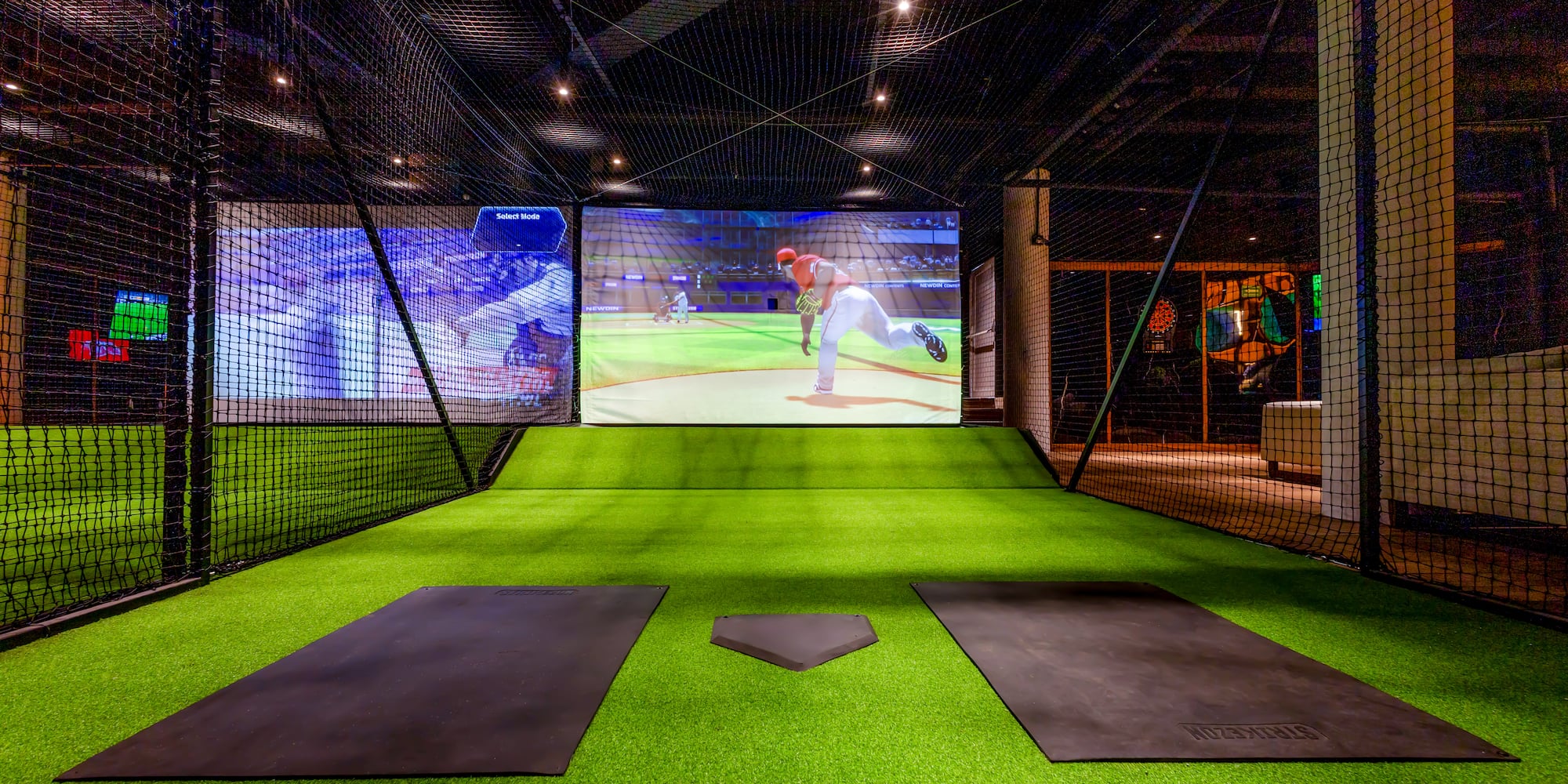 Indoor baseball simulator with green turf, nets, and virtual game screen.