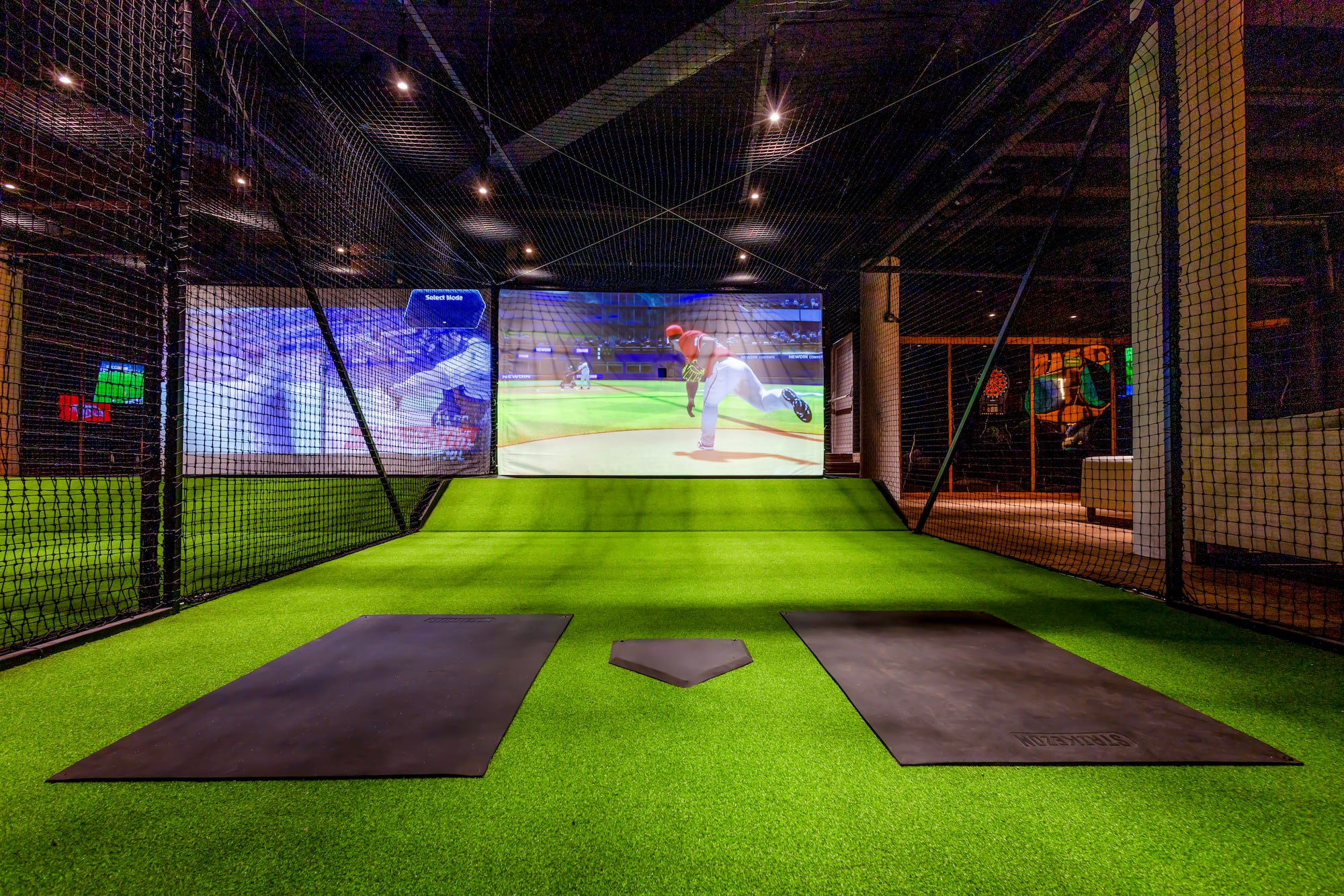 Indoor baseball simulator with green turf, nets, and virtual game screen.