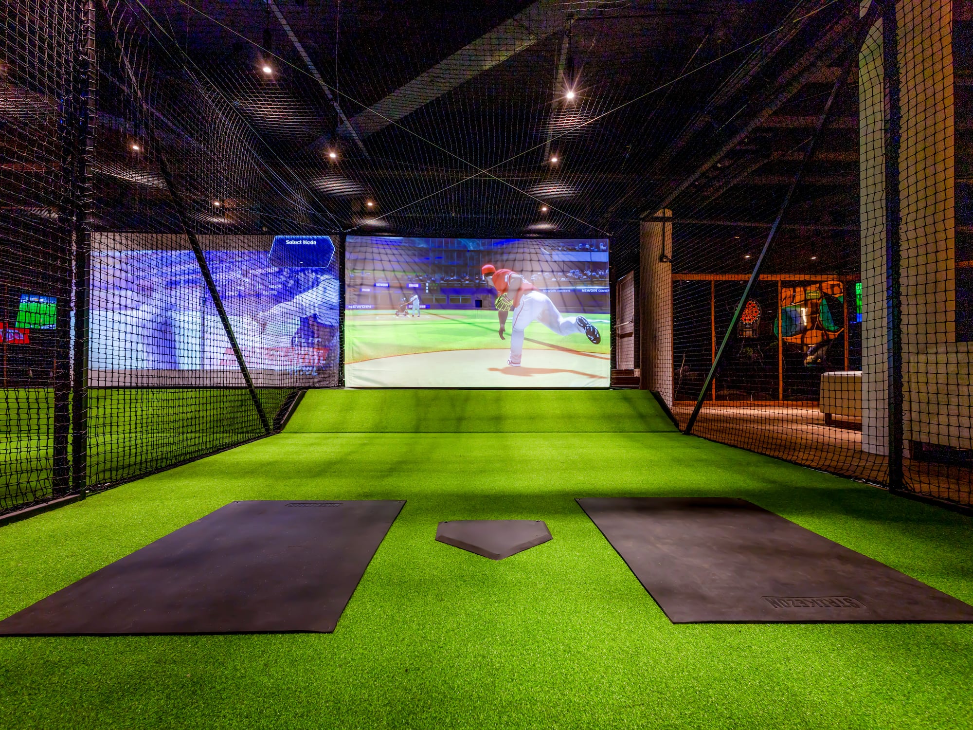 Simulador de béisbol virtual con pantalla, césped artificial y home plate en jaula de bateo.