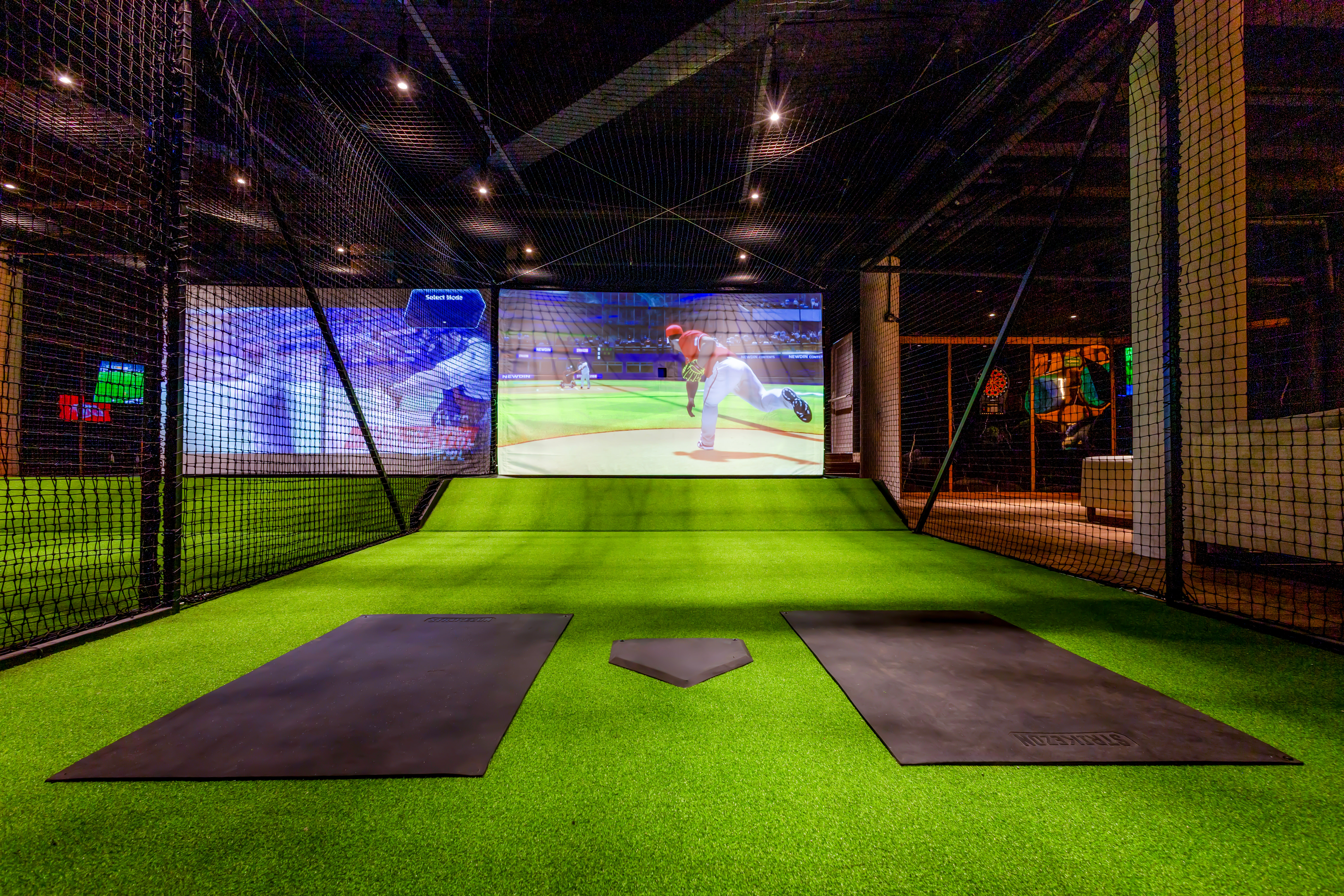 Simulador de béisbol virtual con pantalla, césped artificial y home plate en jaula de bateo.