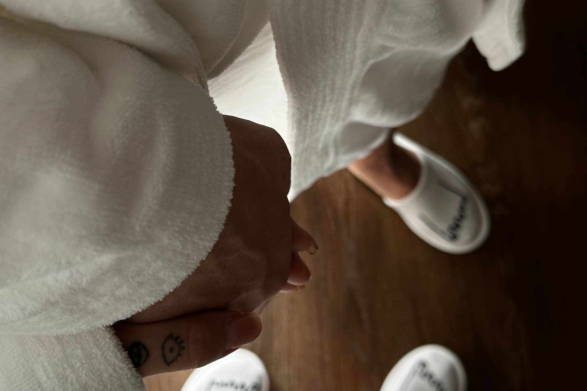 Couple in white bathrobes and hotel slippers holding hands on a wooden floor.