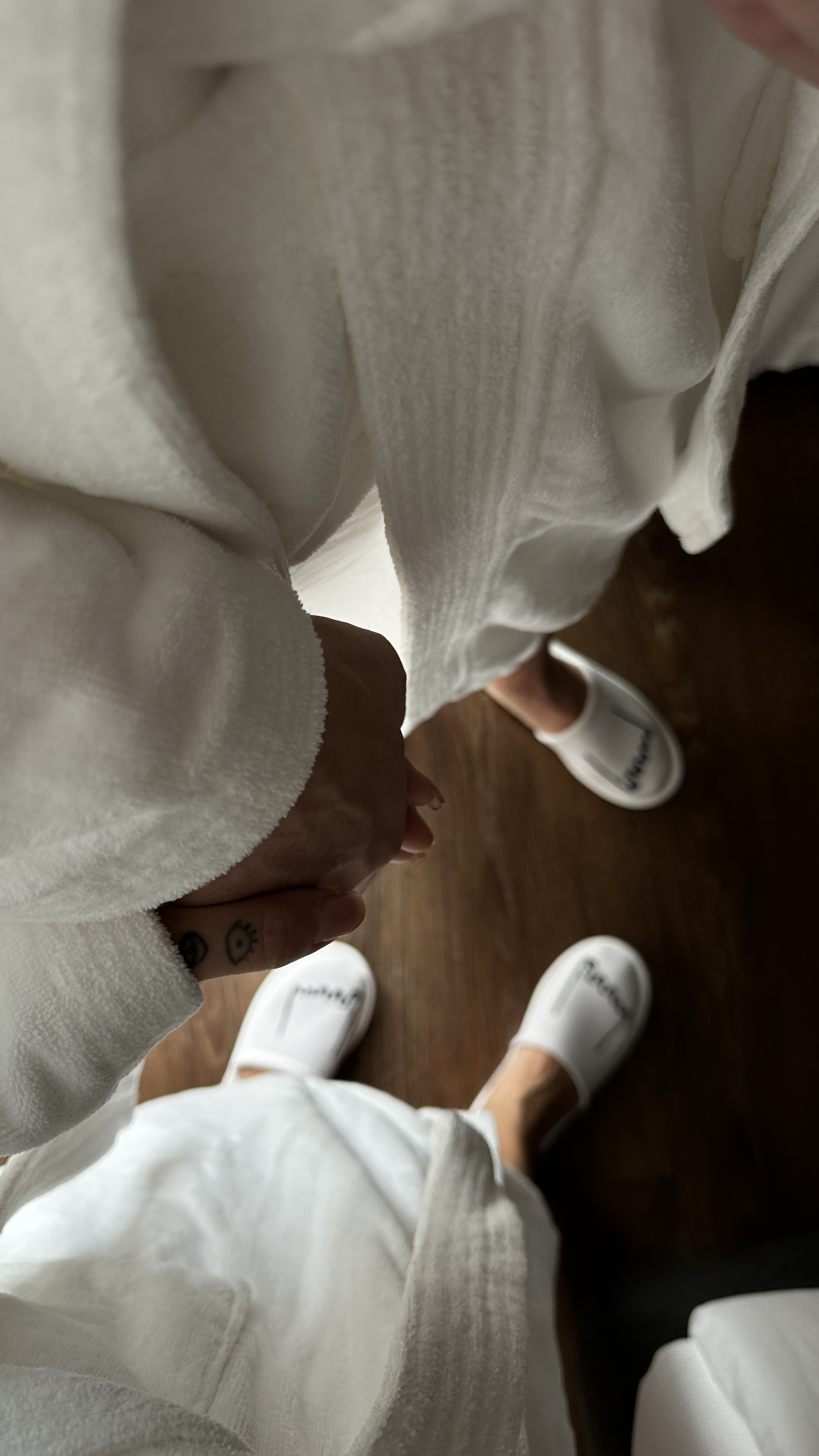 Couple in white bathrobes and hotel slippers holding hands on a wooden floor.