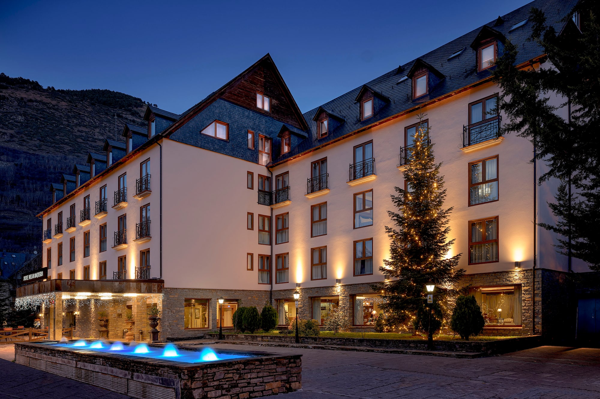 Illuminated hotel exterior at night with Christmas tree and blue fountain.