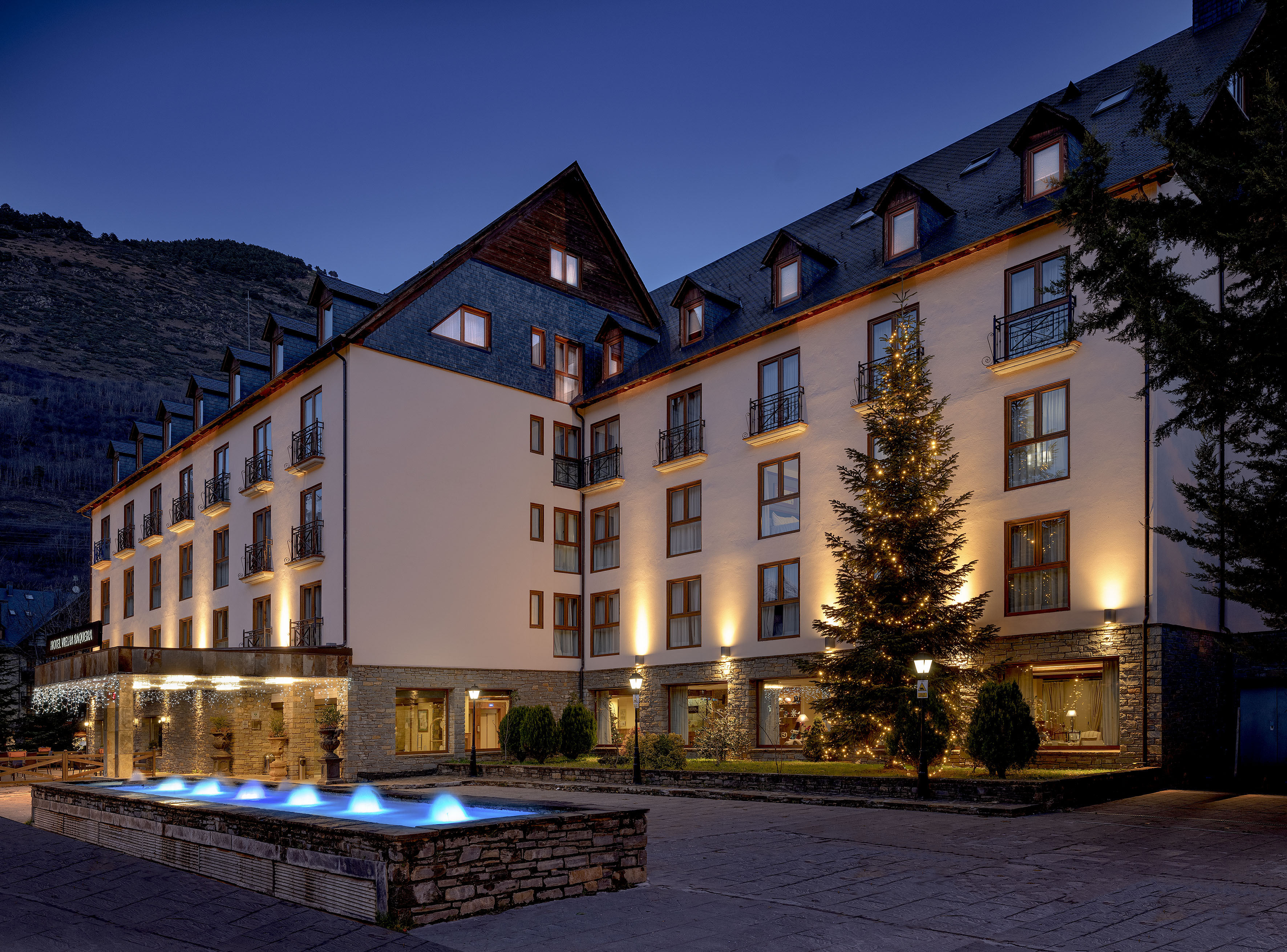Illuminated hotel exterior at night with Christmas tree and blue fountain.