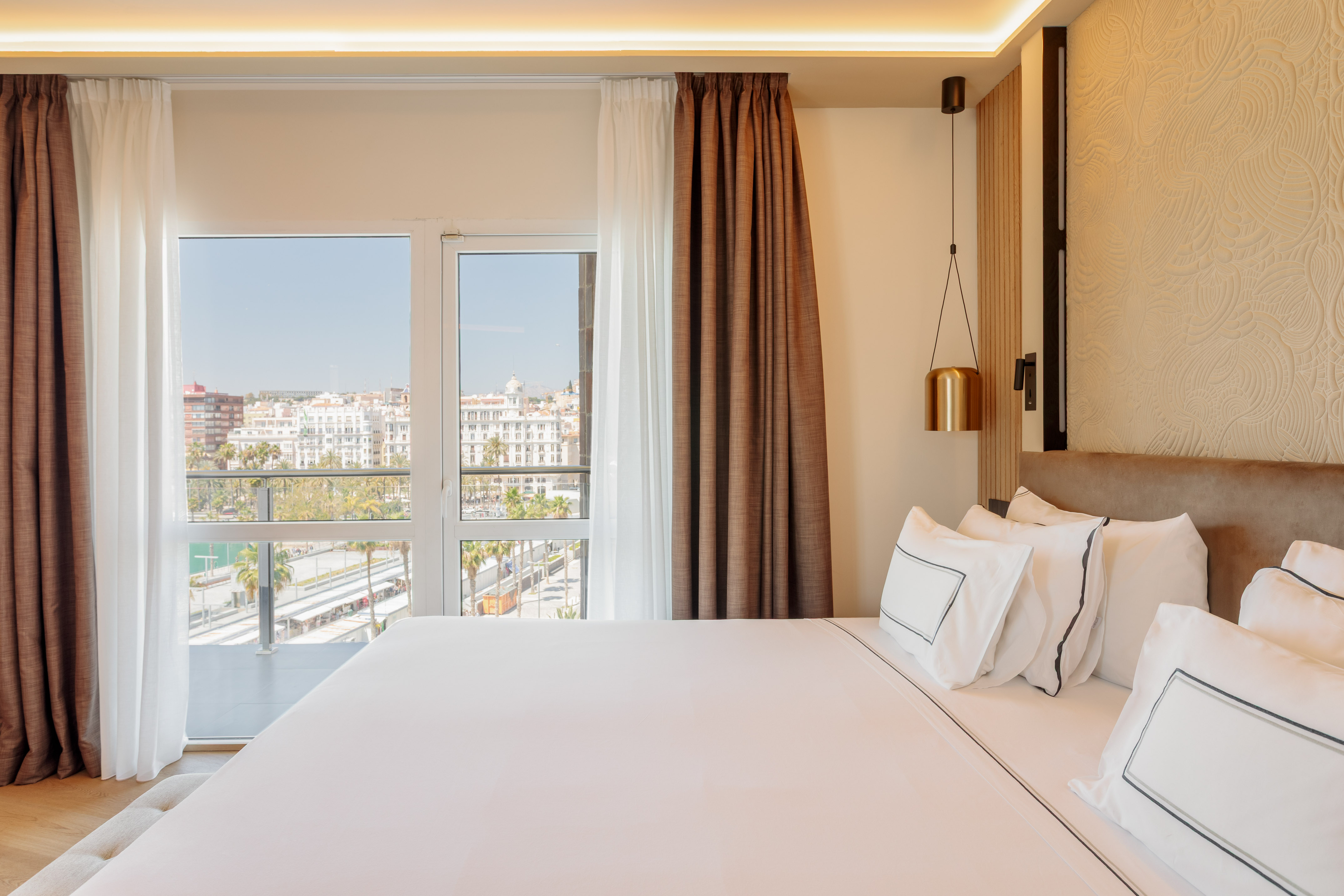 Bright hotel room with white bed, brown curtains, and city-port view.