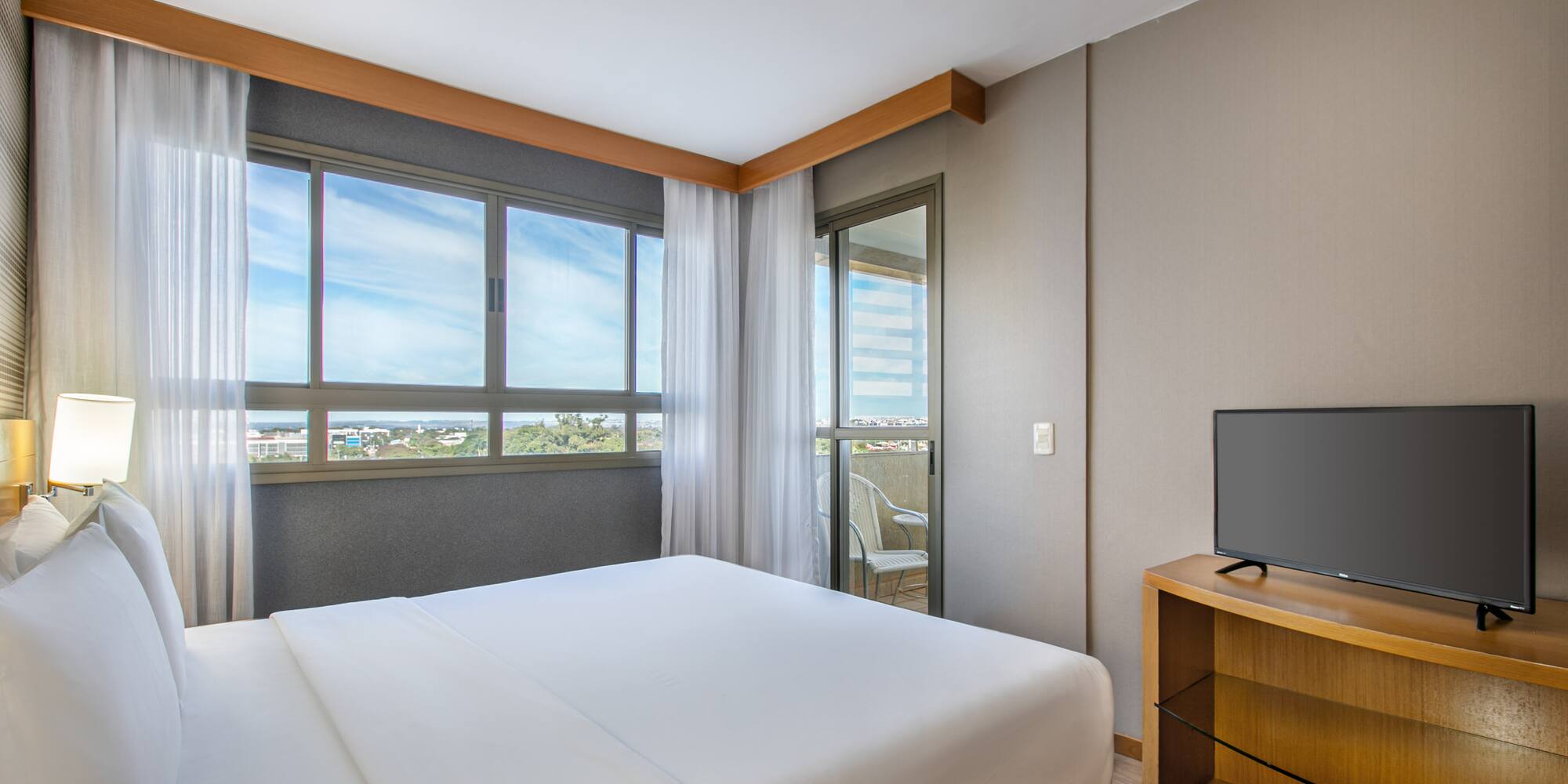 Modern hotel bedroom with king bed, balcony, city view, and TV.
