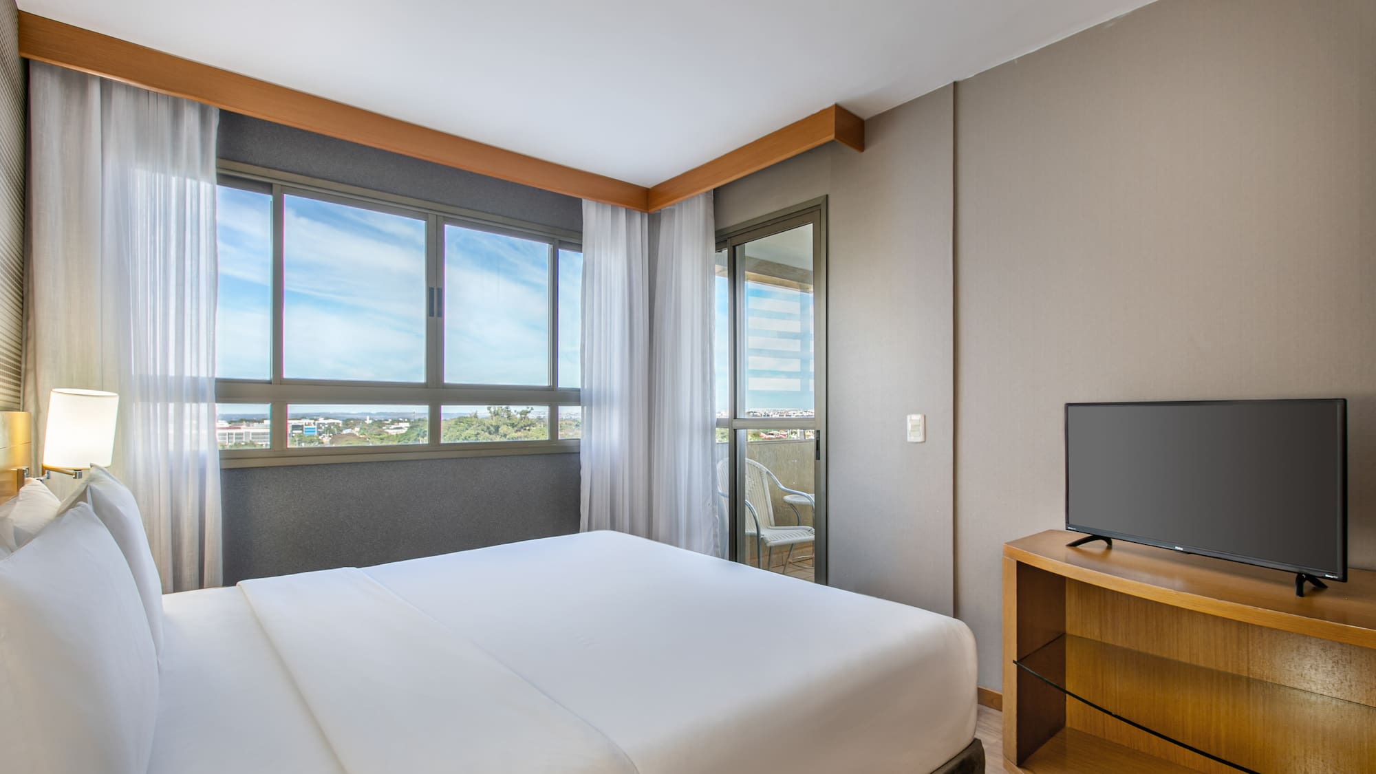 Modern hotel bedroom with king bed, balcony, city view, and TV.