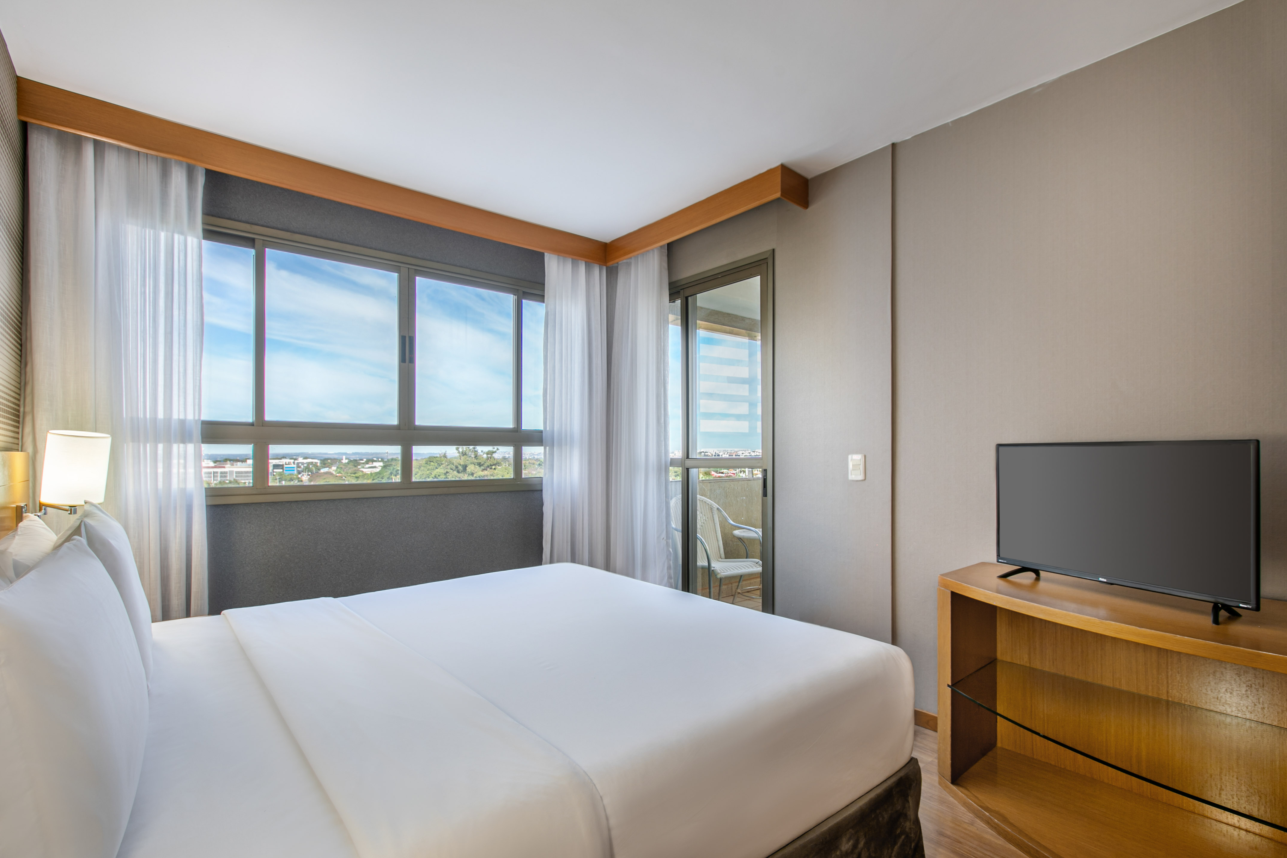 Modern hotel bedroom with king bed, balcony, city view, and TV.