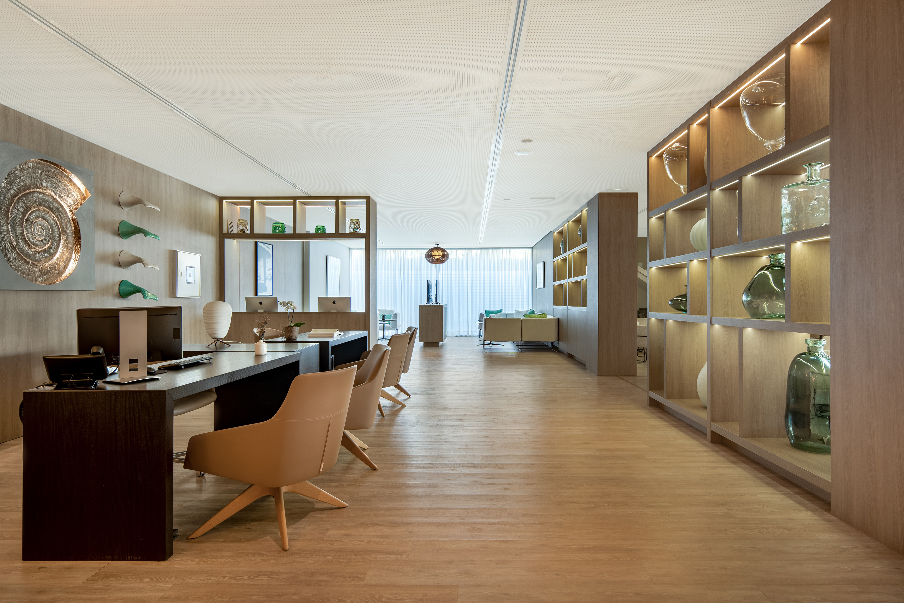 Modern office reception with wooden desk, tan chairs, and illuminated shelving.