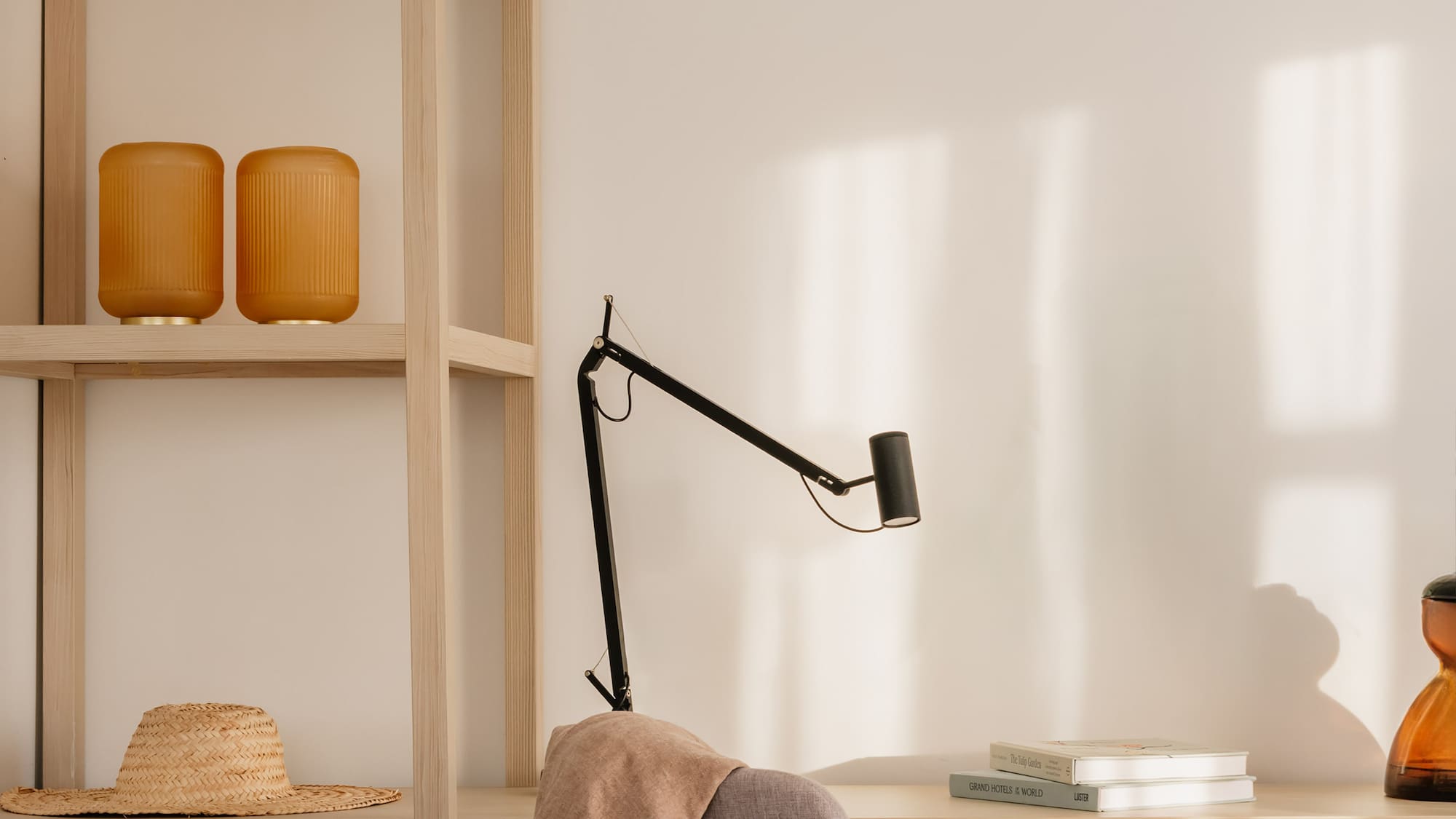 Sunlit home office with light wood desk, grey chair, black lamp, and shelf.