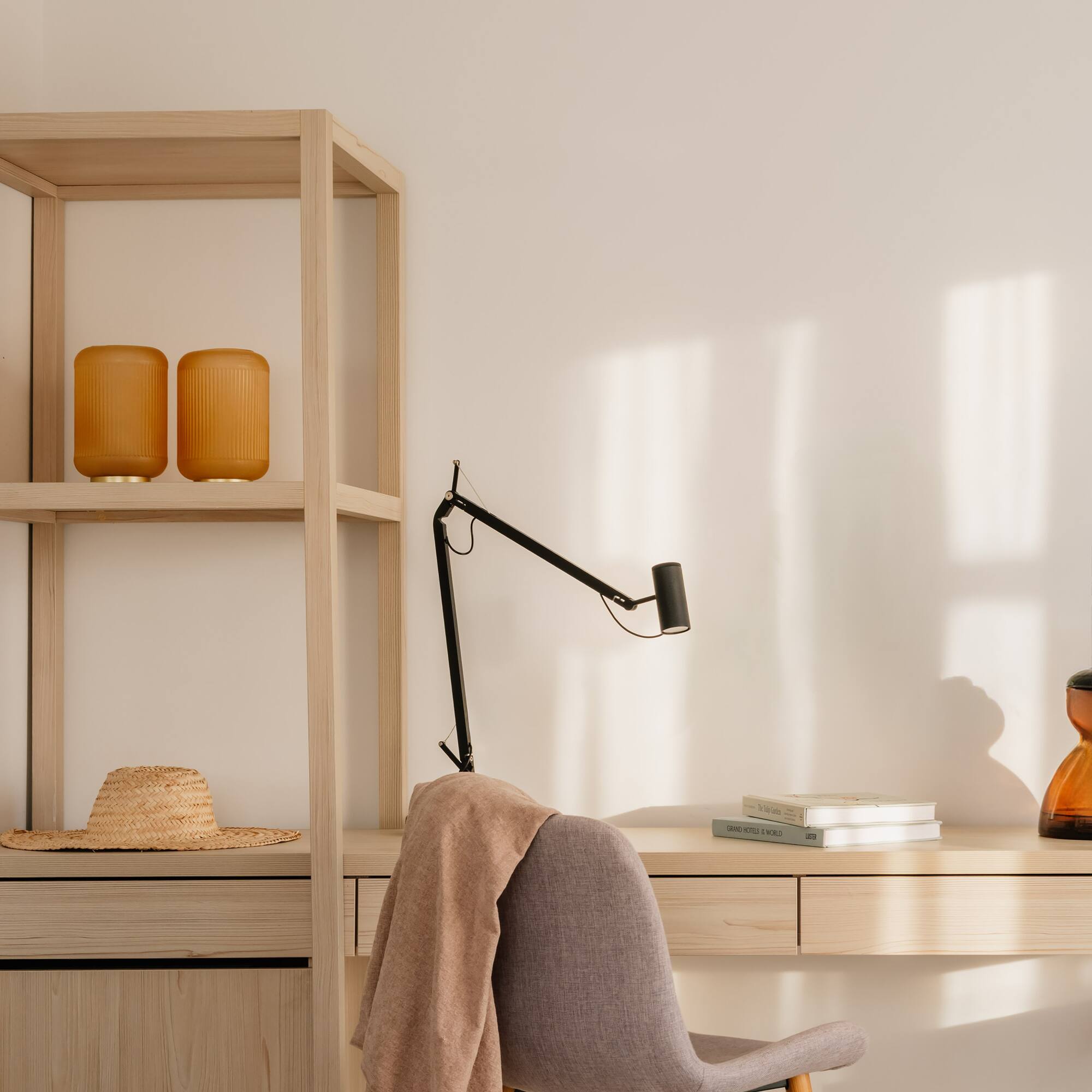 Sunlit home office with light wood desk, grey chair, black lamp, and shelf.