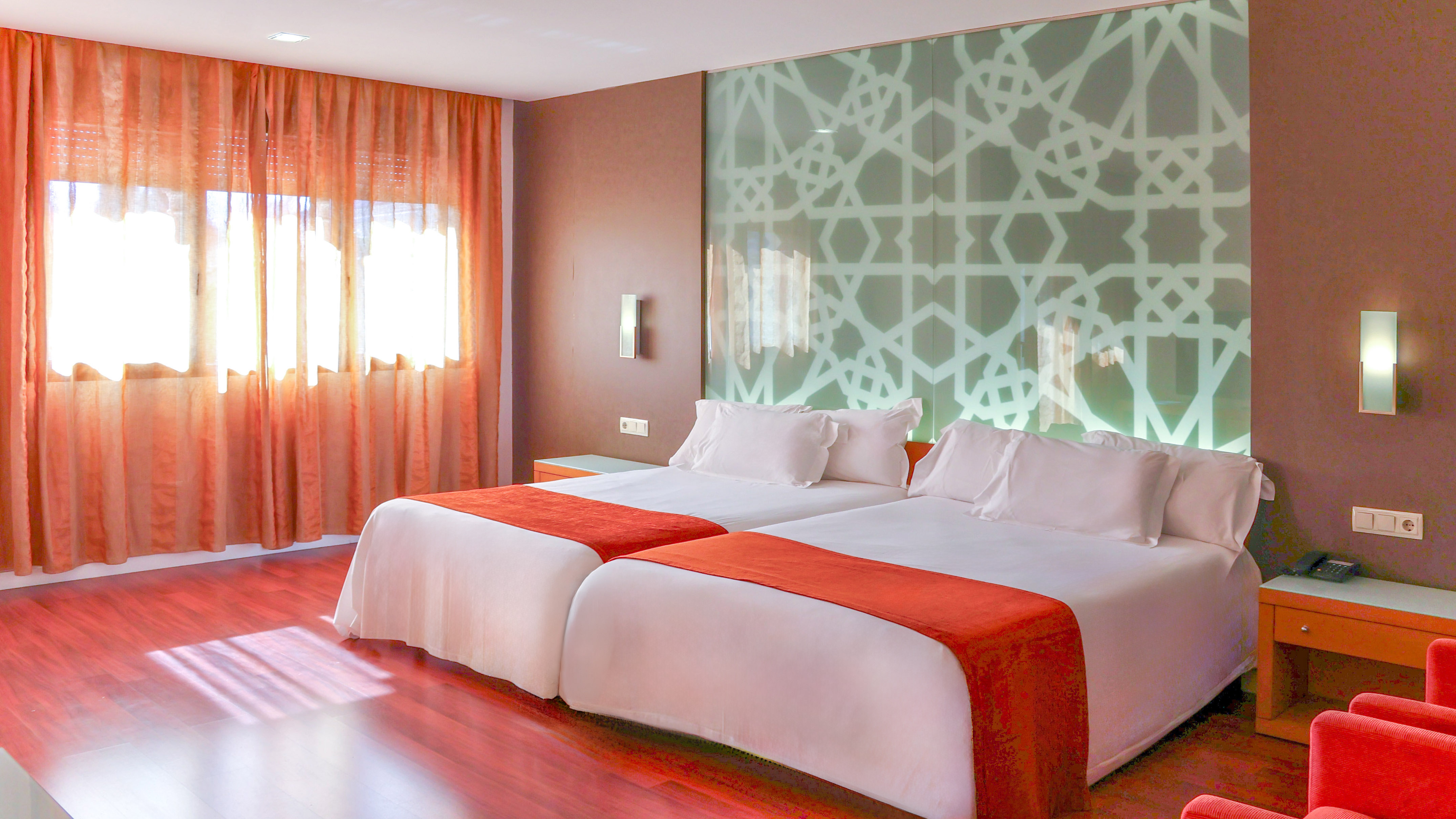 Modern hotel room with two twin beds, orange decor, and geometric patterned wall.