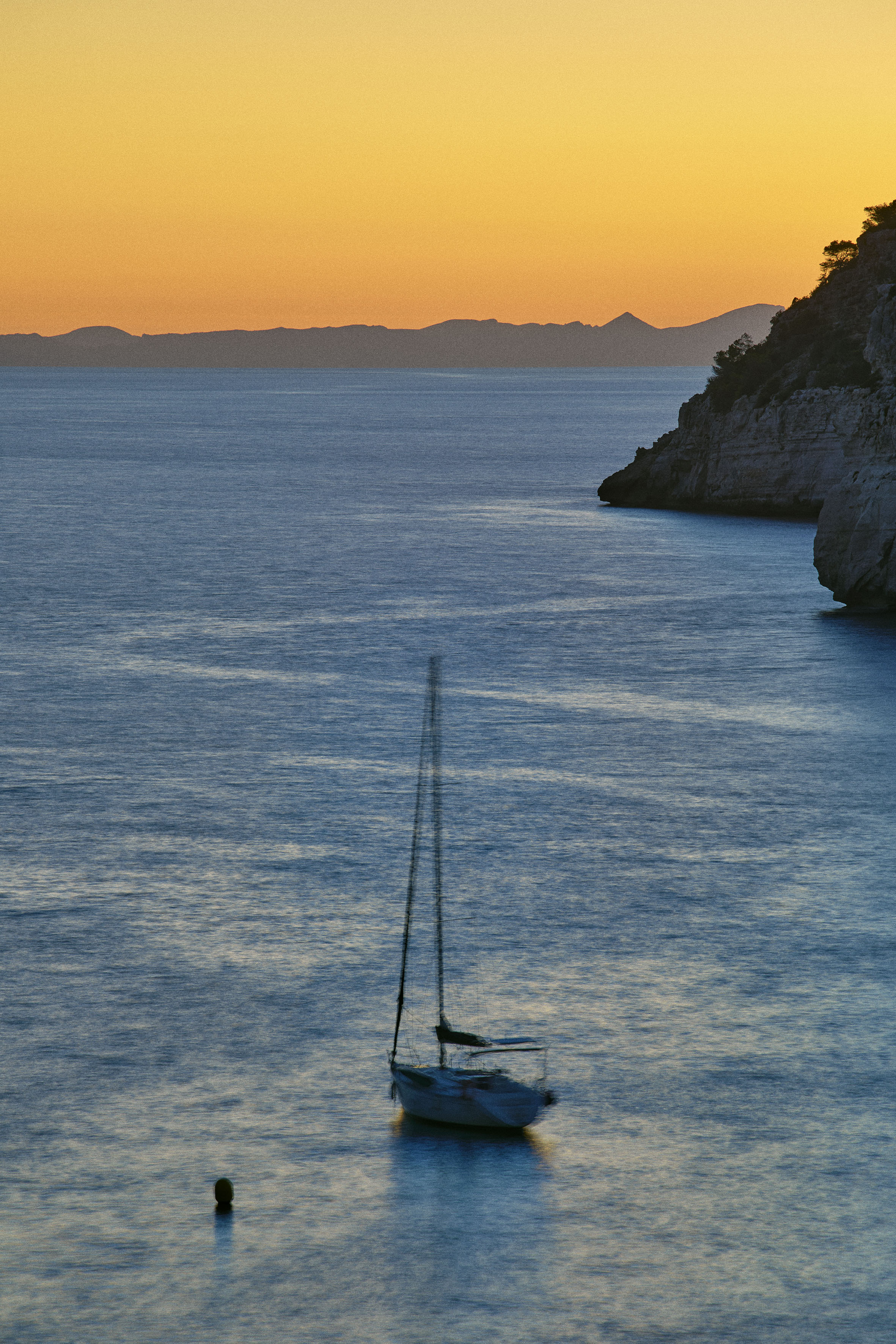 Blurred sailboat on blue water, orange sunset sky, coastal cliffs.
