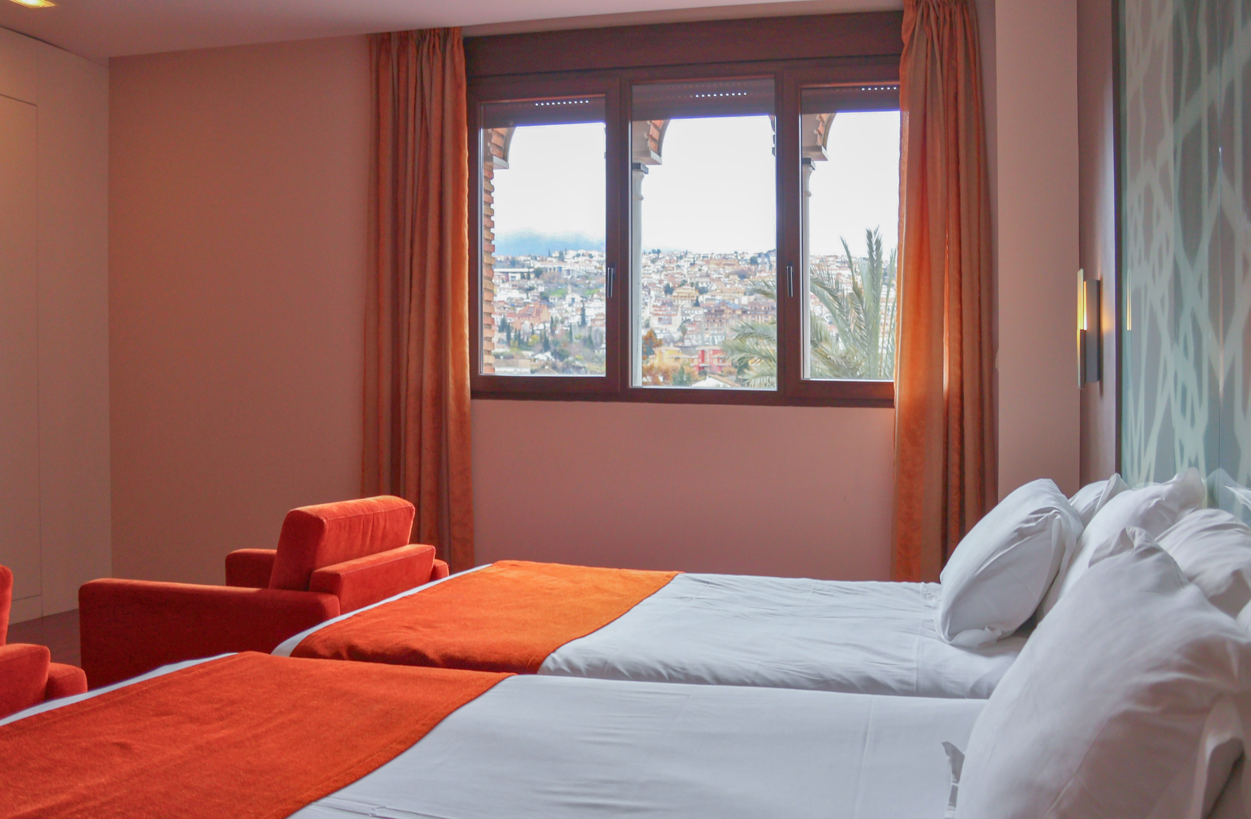 Hotel room with twin beds, orange throws, red armchairs, and a scenic city view.