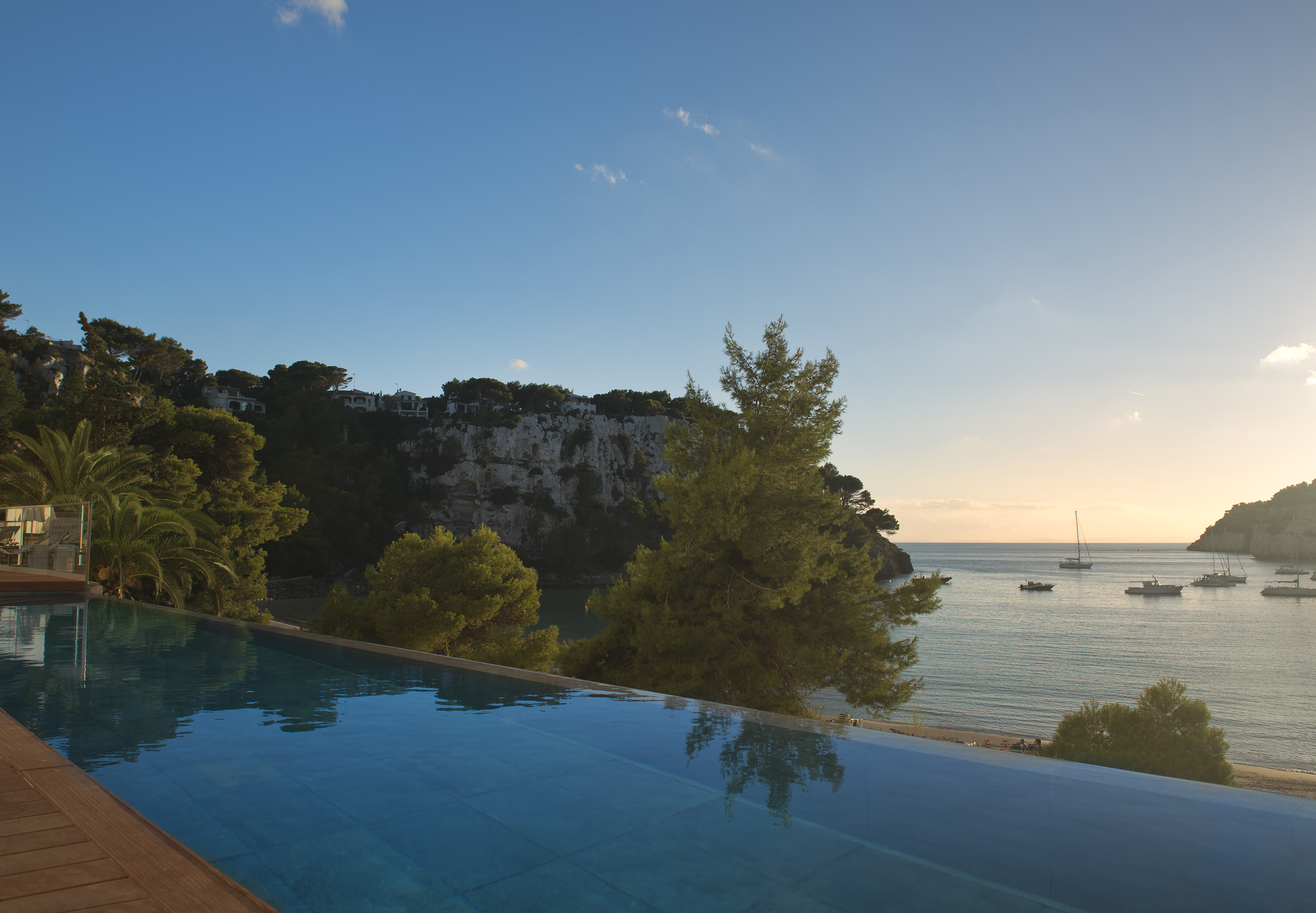 Infinity pool with ocean view, cliffside villas, and boats at sunset.