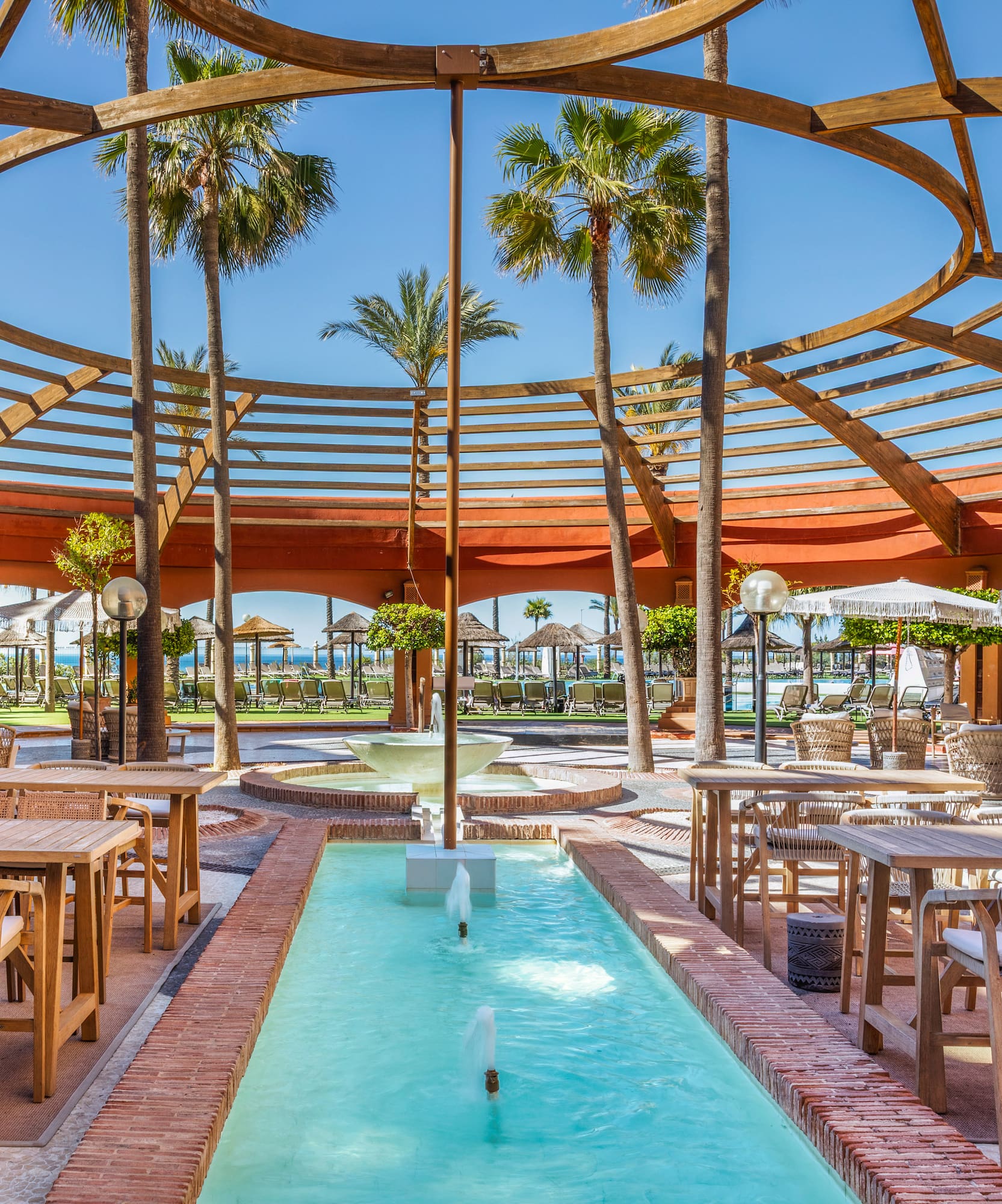 Outdoor resort dining under circular pergola, with palm trees and a long fountain.