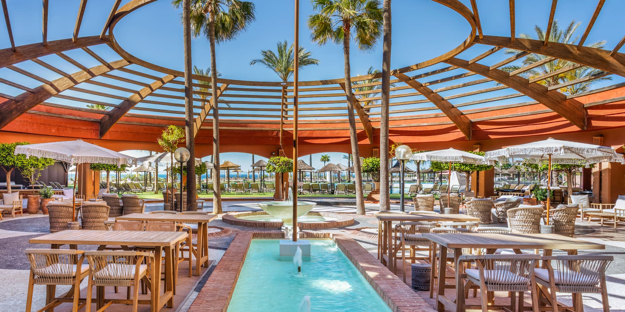 Resort outdoor lounge with circular pergola, palm trees, and fountain.