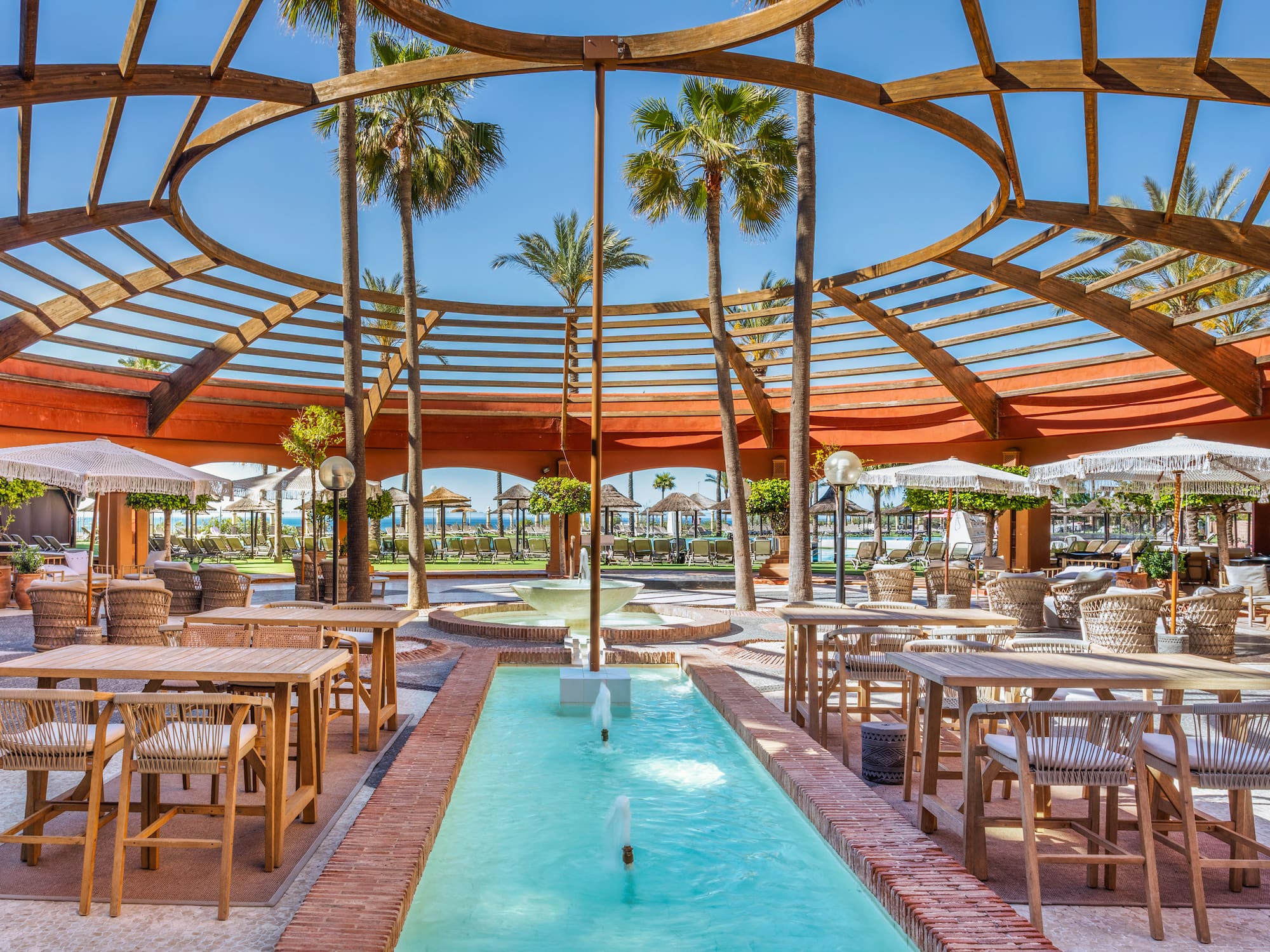 Resort outdoor lounge with circular pergola, palm trees, and fountain.