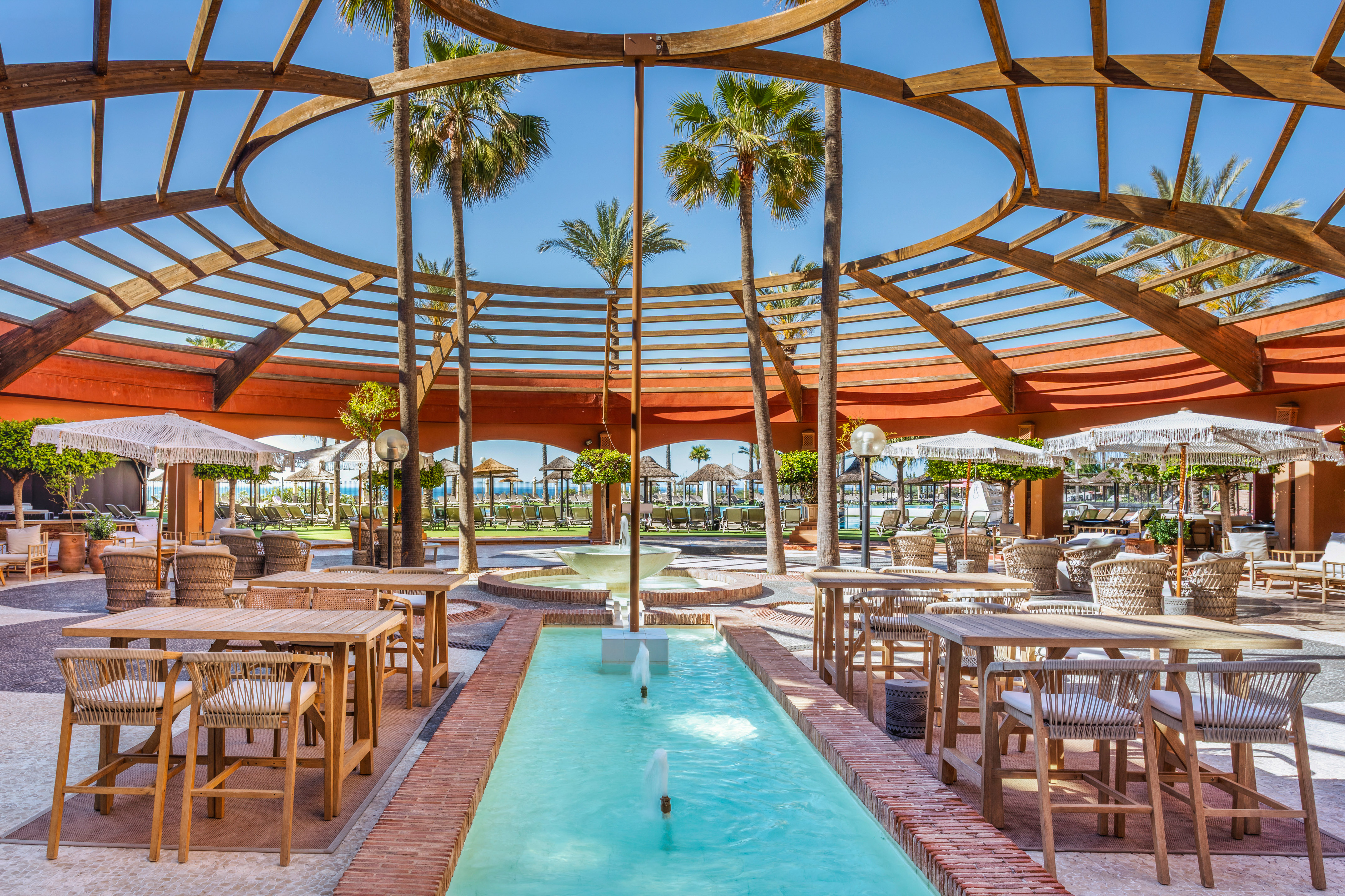 Resort outdoor lounge with circular pergola, palm trees, and fountain.