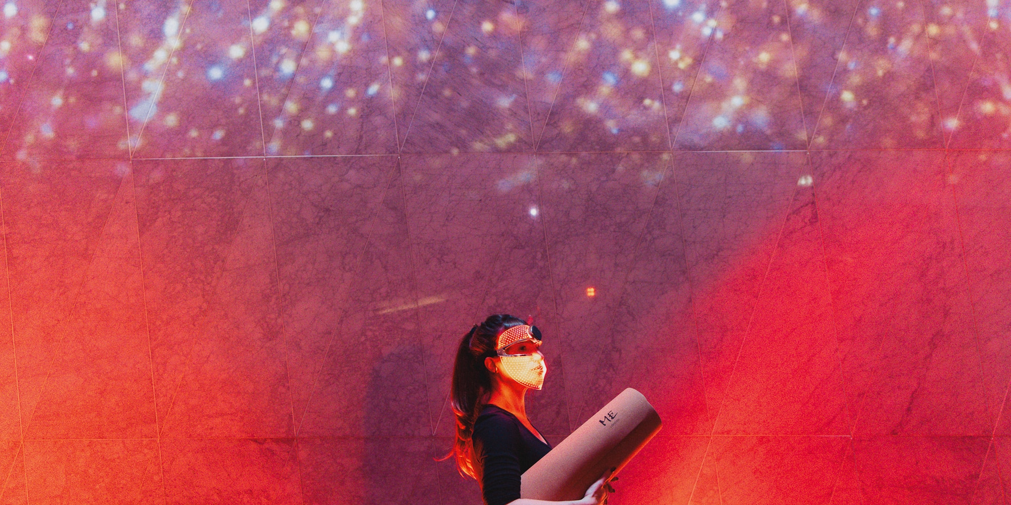 Woman in glowing LED mask holding yoga mat against sparkling cosmic wall.